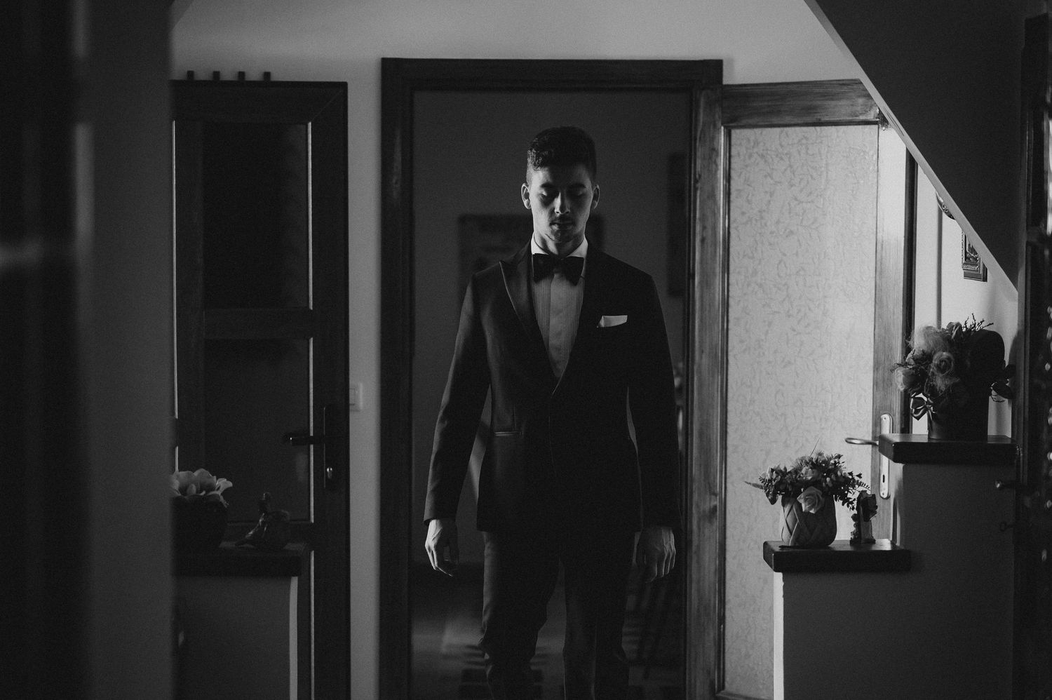 Groom in black tuxedo standing in doorway in dramatic black and white photo.