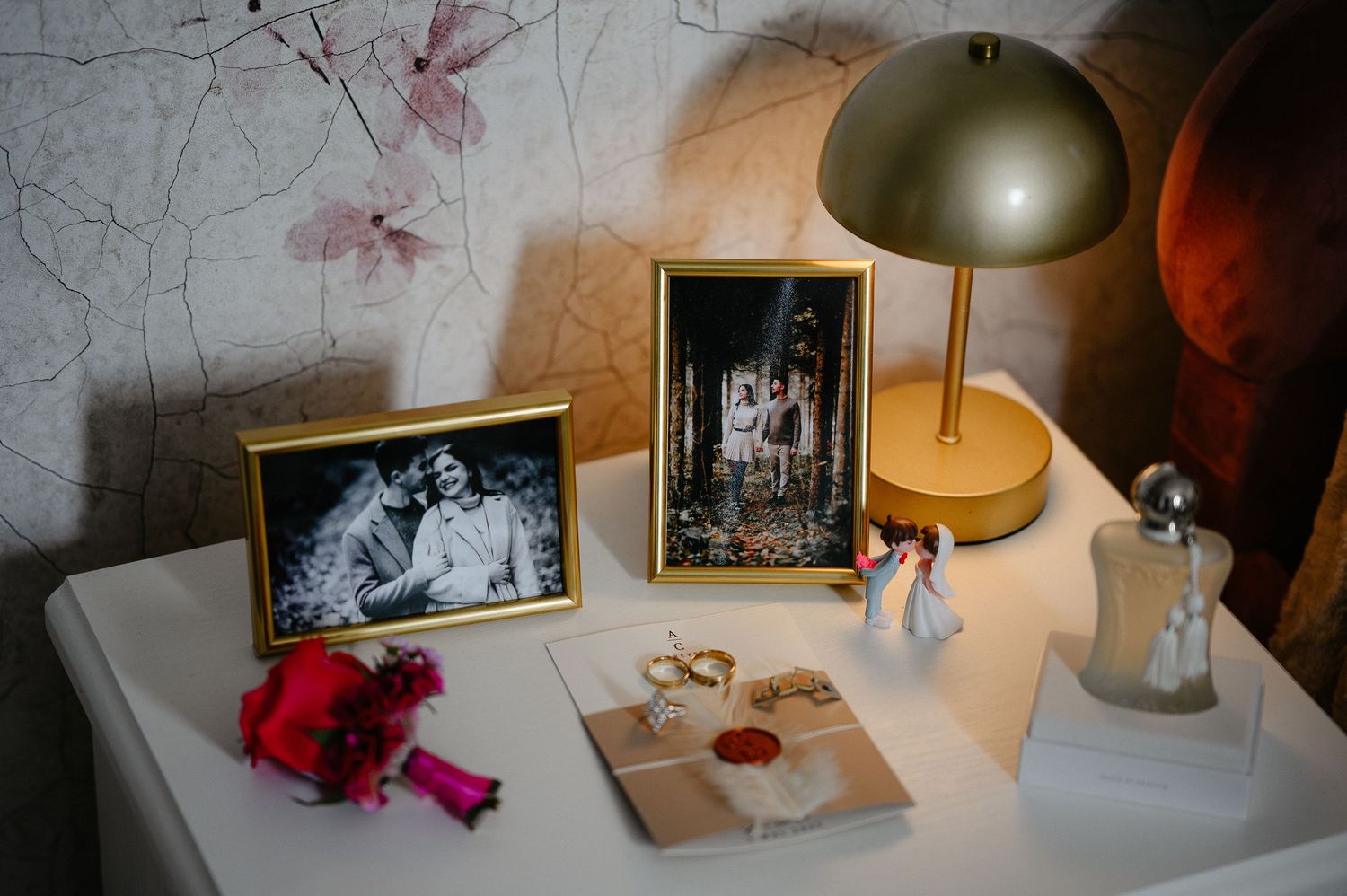 Wedding details flat lay with framed couple photos, gold rings, bouquet, and lamp on white nightstand.
