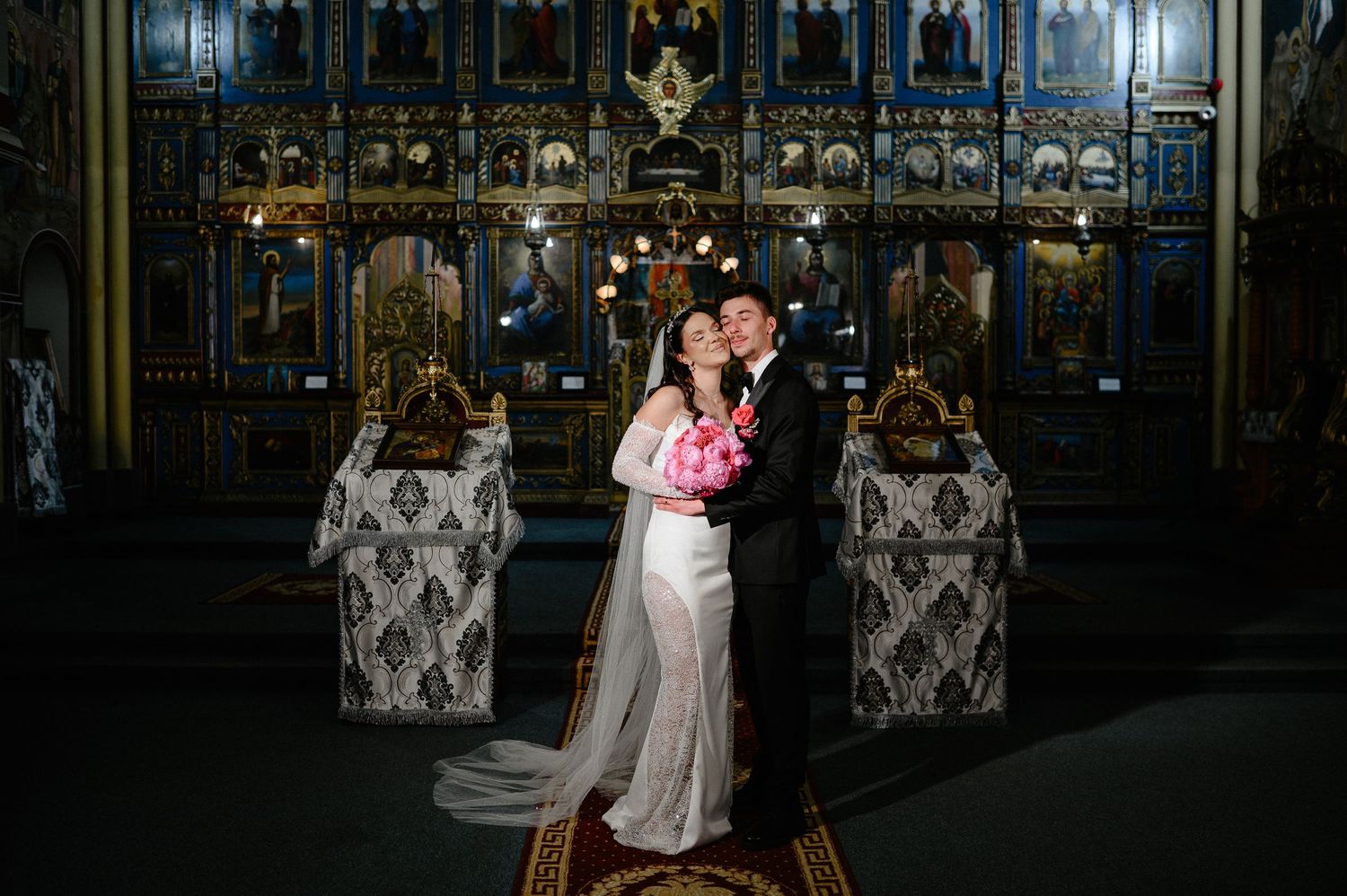 Bride and groom share a romantic embrace before the ornate golden iconostasis of an Orthodox church.