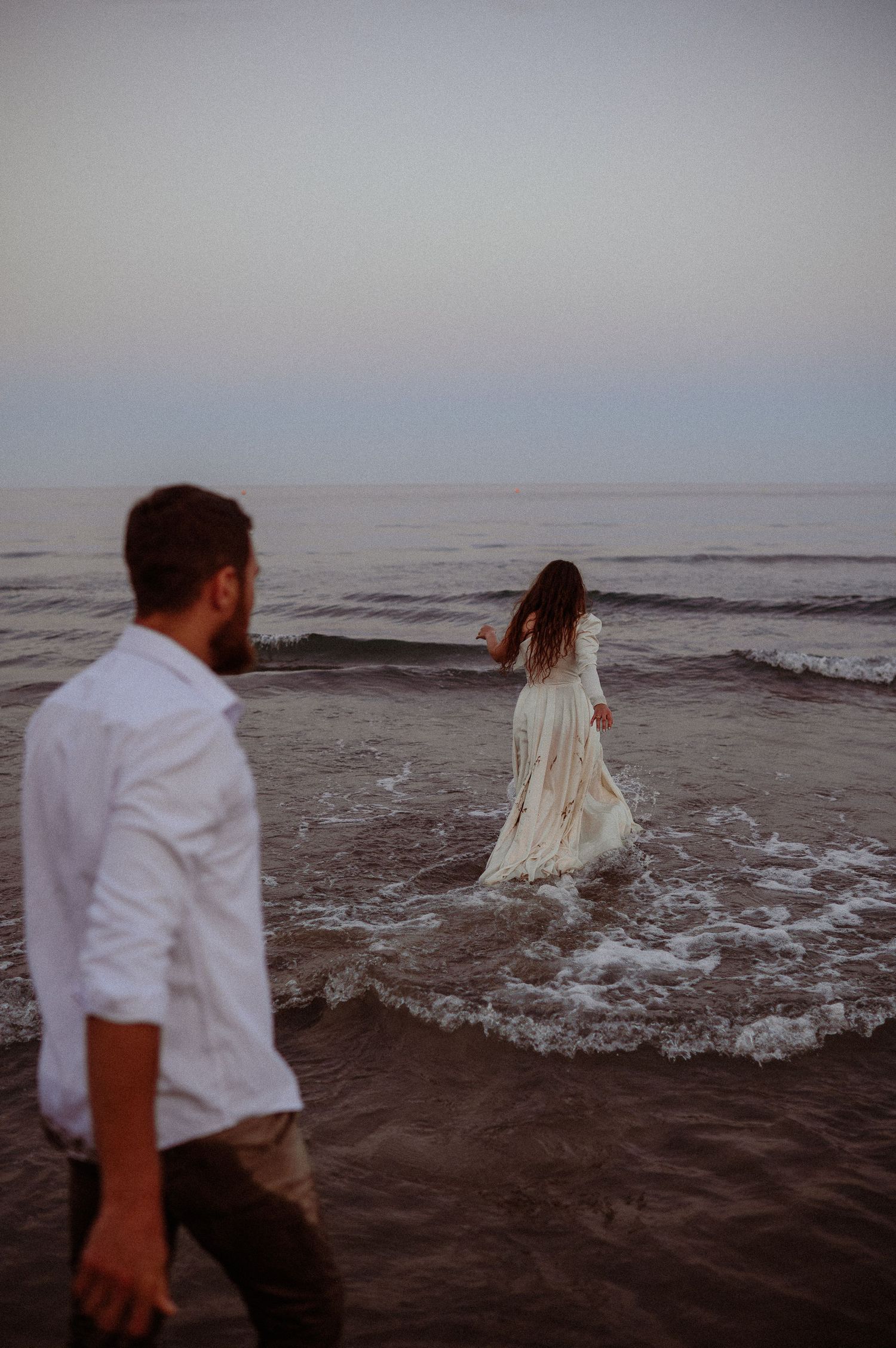 Bride in flowing white dress wading in ocean waves at sunset while groom watches from the shore.