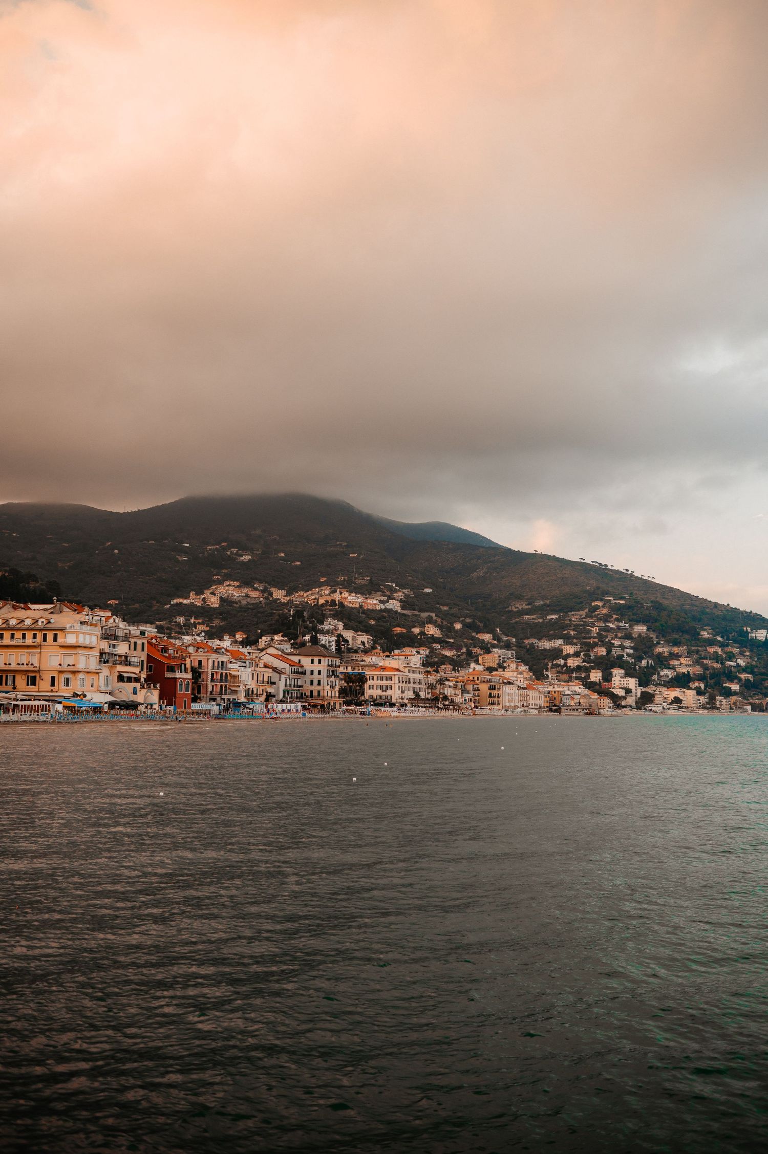 Coastal Italian town with colorful buildings nestled at the foot of misty green mountains at sunset.