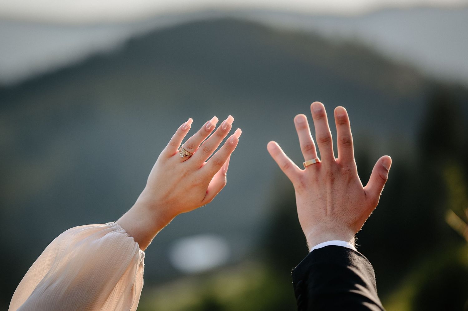Close-up of couple raising hands showing wedding rings against a blurred mountain backdrop.