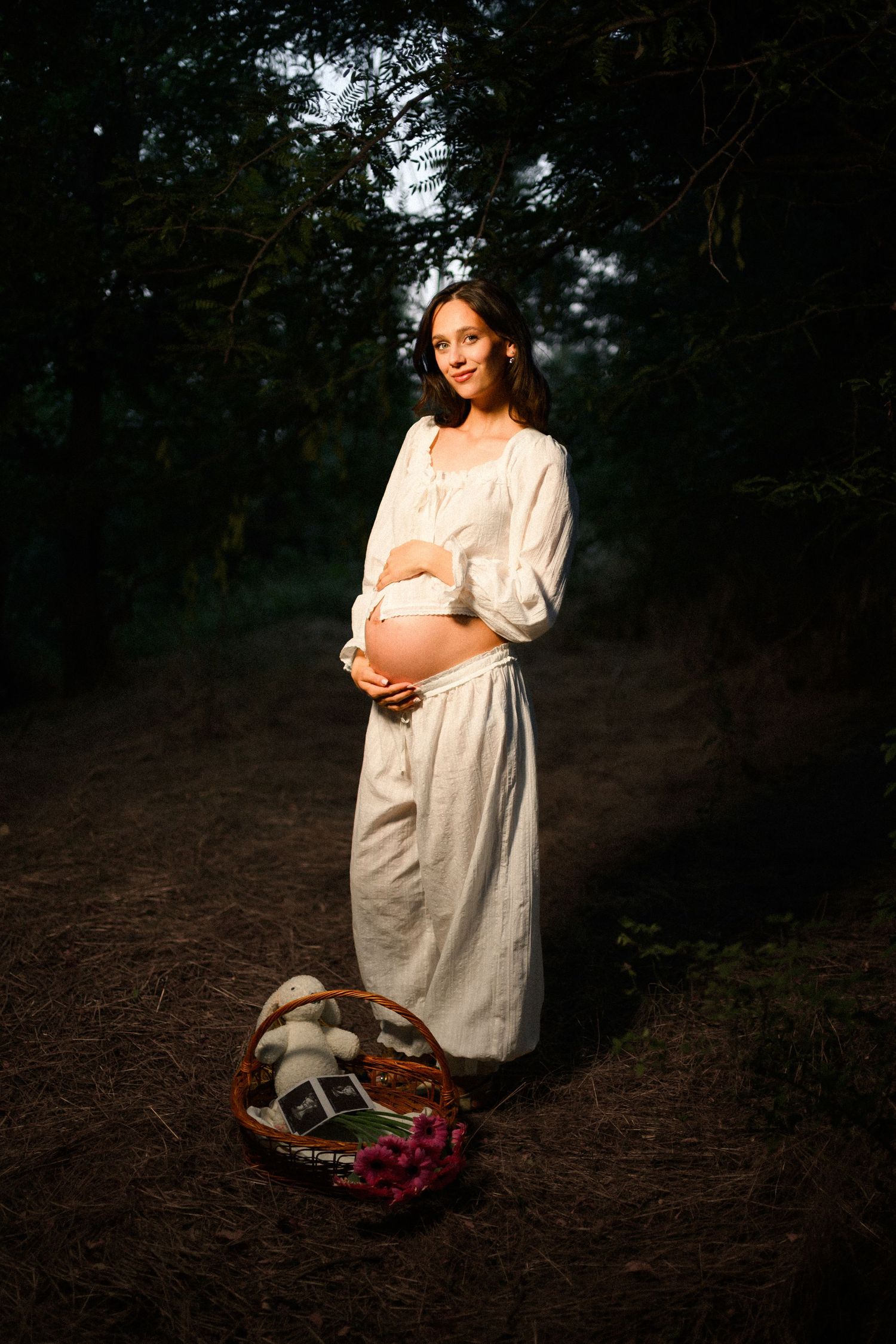 Pregnant woman in white dress cradling belly in forest, with wicker basket of flowers at her feet.