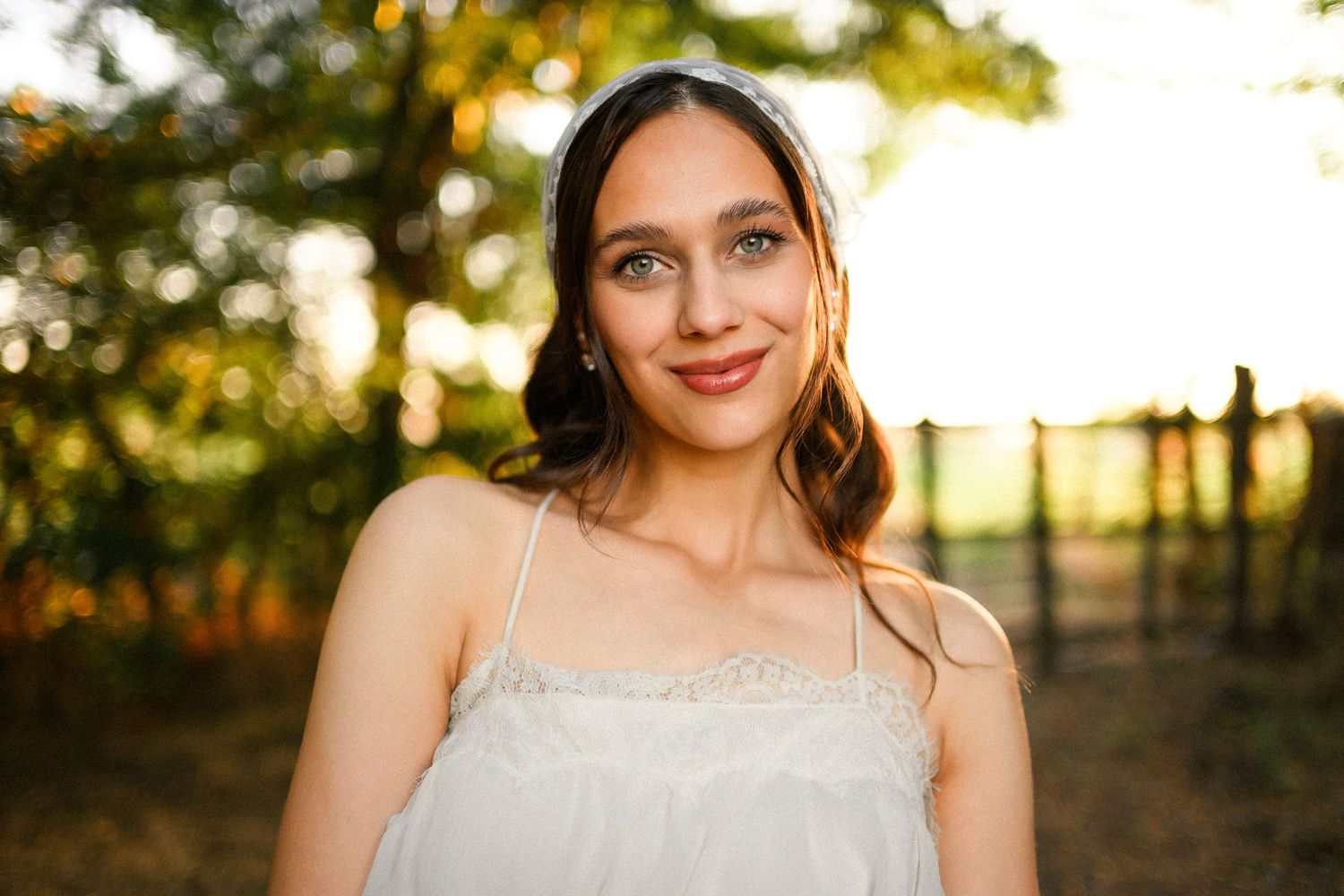 Young woman in white sundress smiling outdoors at golden hour with bokeh trees in background.