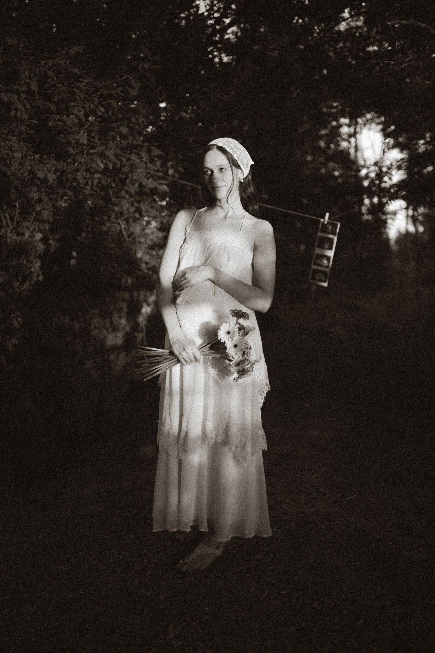 Black and white photo of a woman in a white dress holding flowers and a ladder in a dark forest setting.