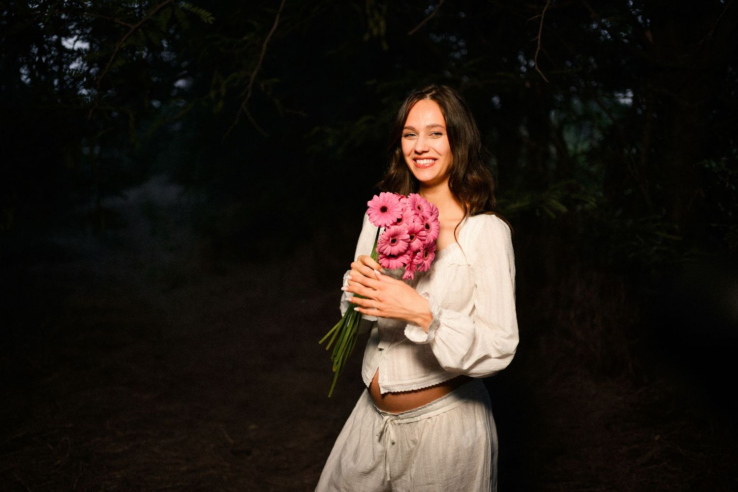 Smiling young woman holding pink peony bouquet outdoors in dark forest setting, wearing white dress.