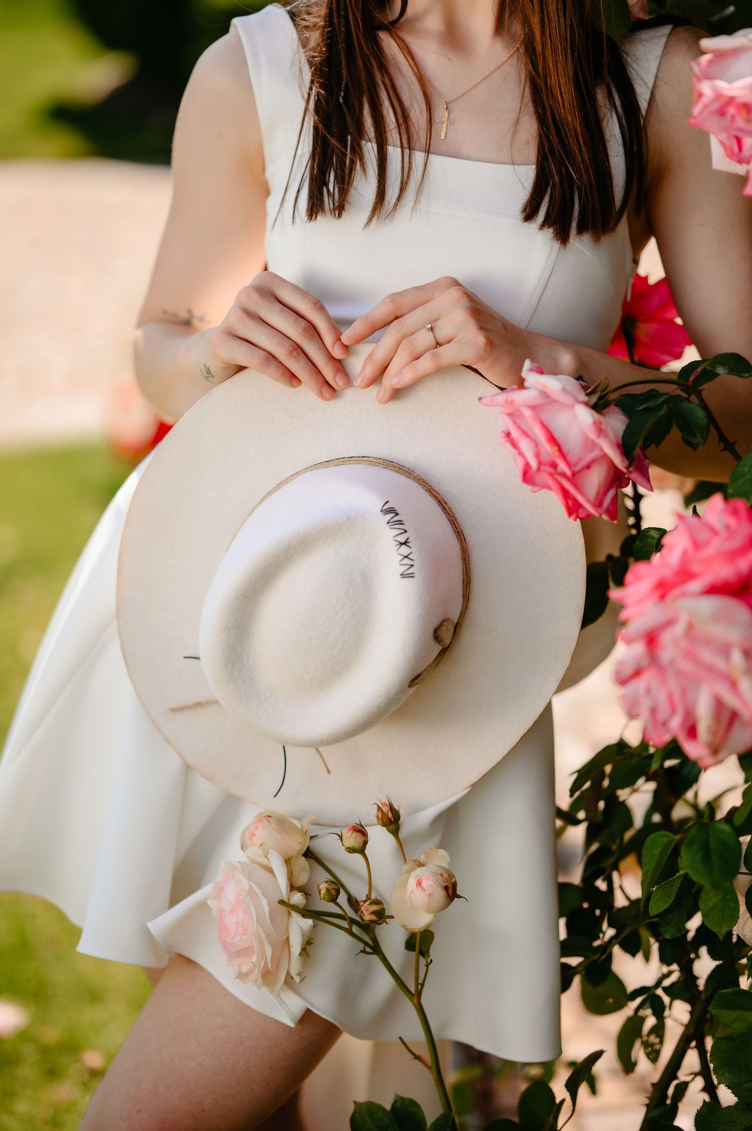 Woman in white dress holding a cream wide-brim hat near pink roses in a garden.