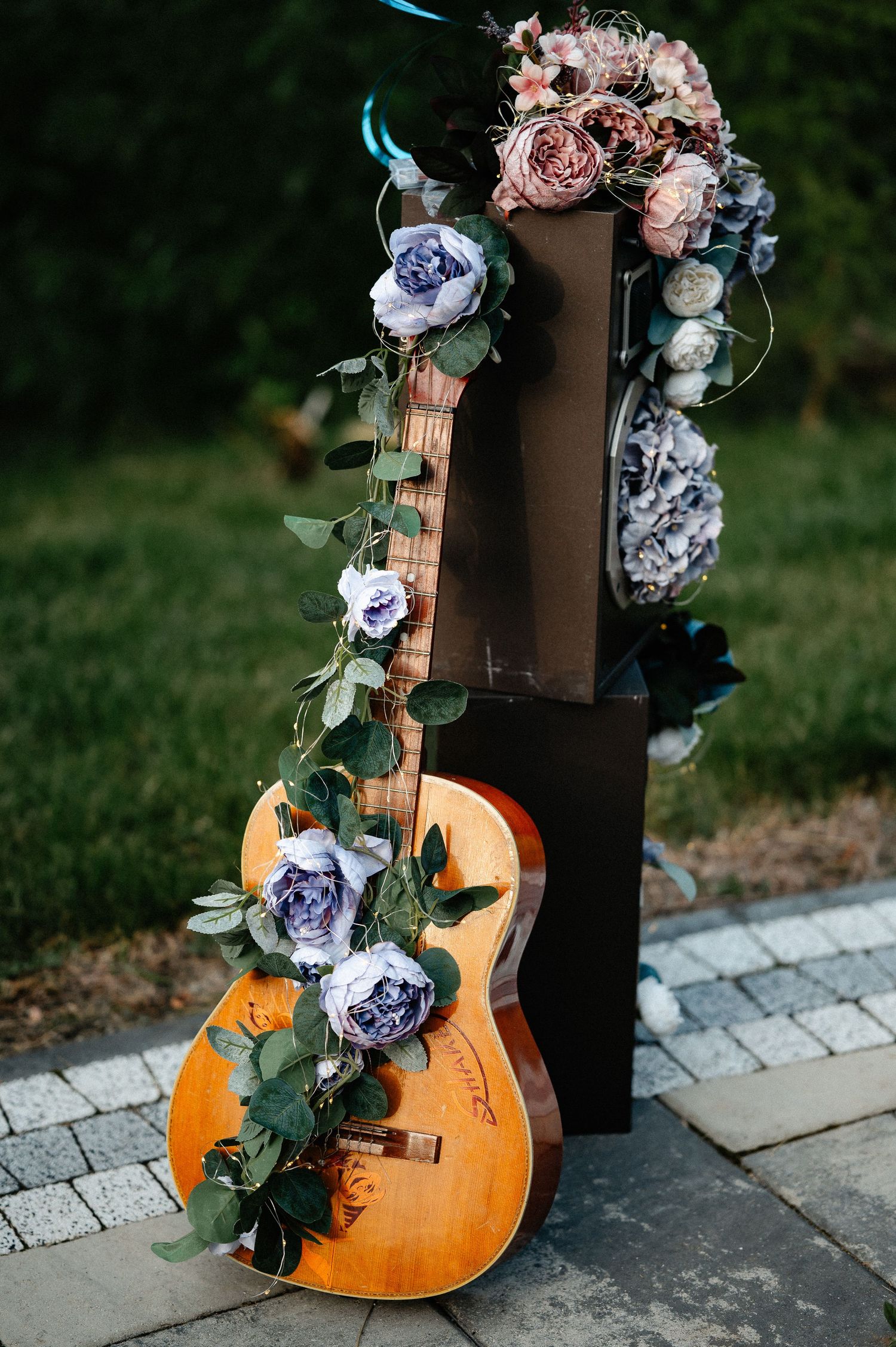 Acoustic guitar decorated with purple roses and eucalyptus garland leaning against dark wooden stand.