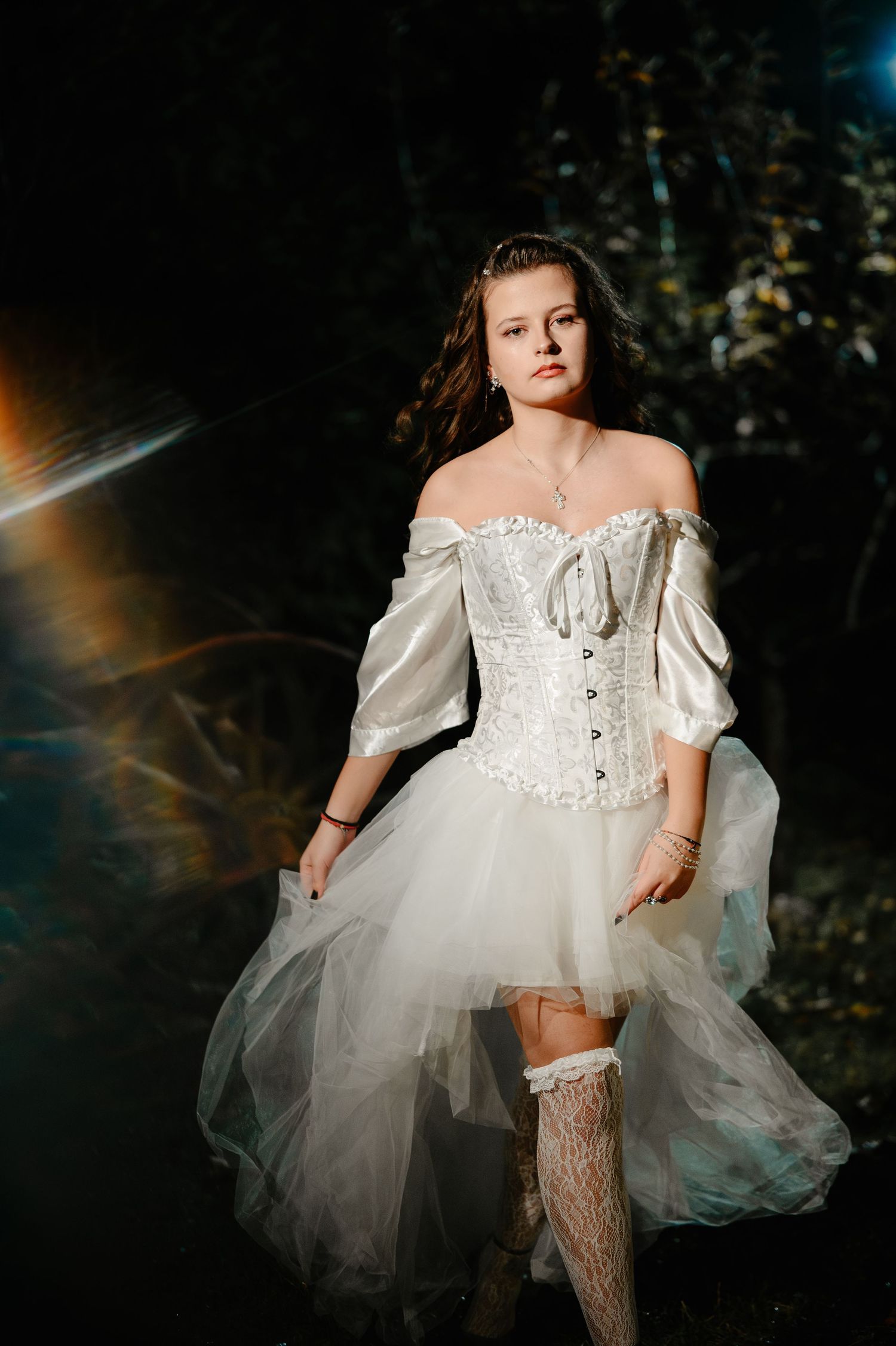 Young woman in white corset dress and lace stockings walks through dark forest with dramatic lighting.