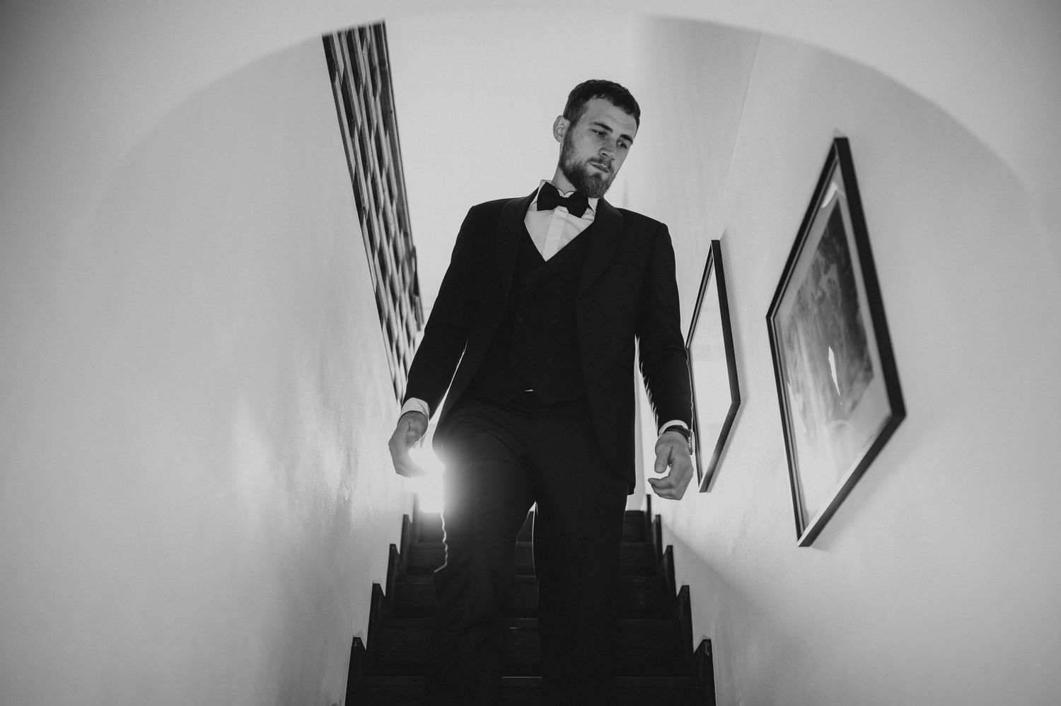 Black and white dramatic shot of a groom in a tuxedo descending a staircase with arched ceiling.