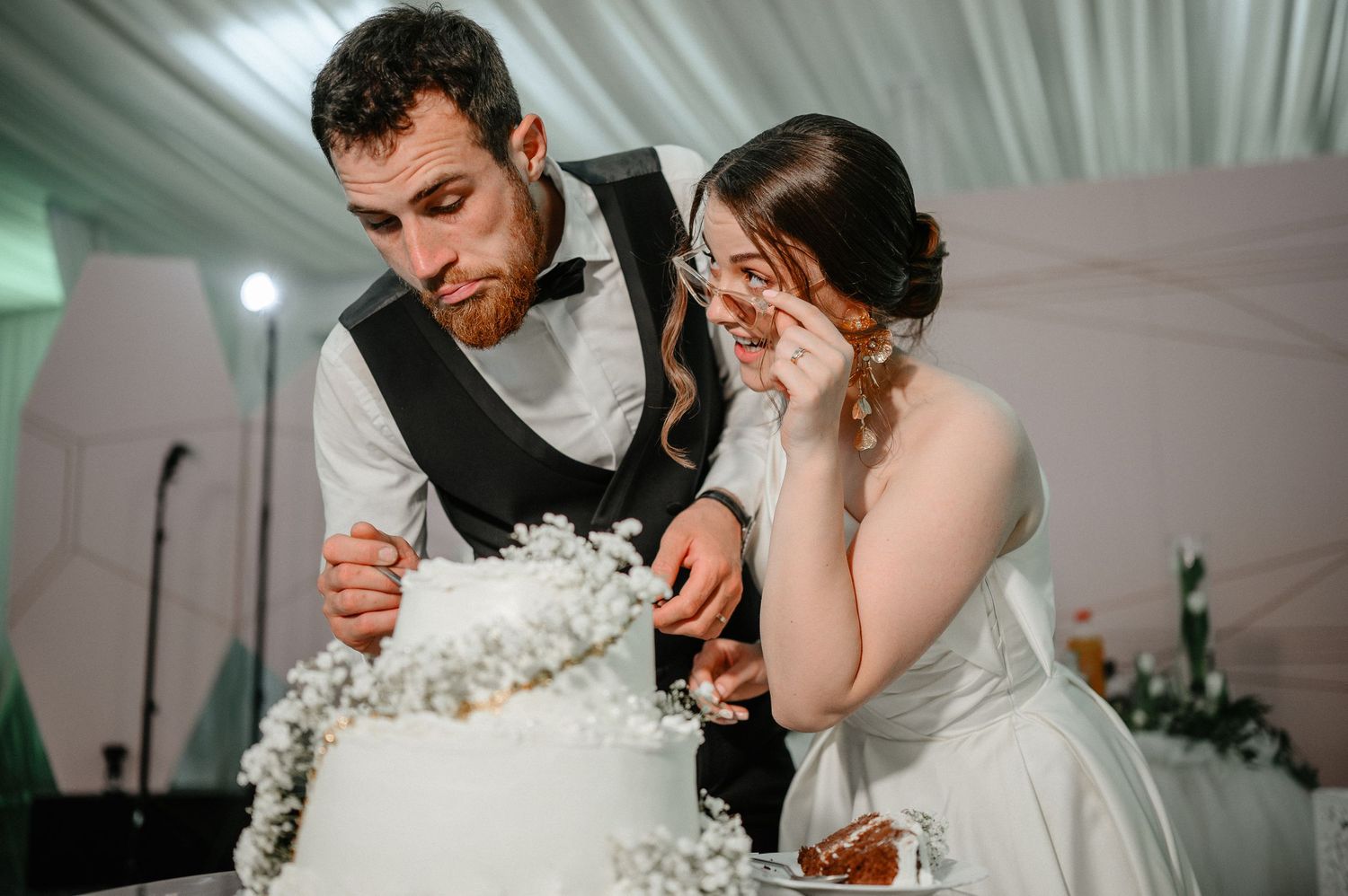 Newlyweds carefully cutting elegant white wedding cake adorned with delicate white floral decorations.