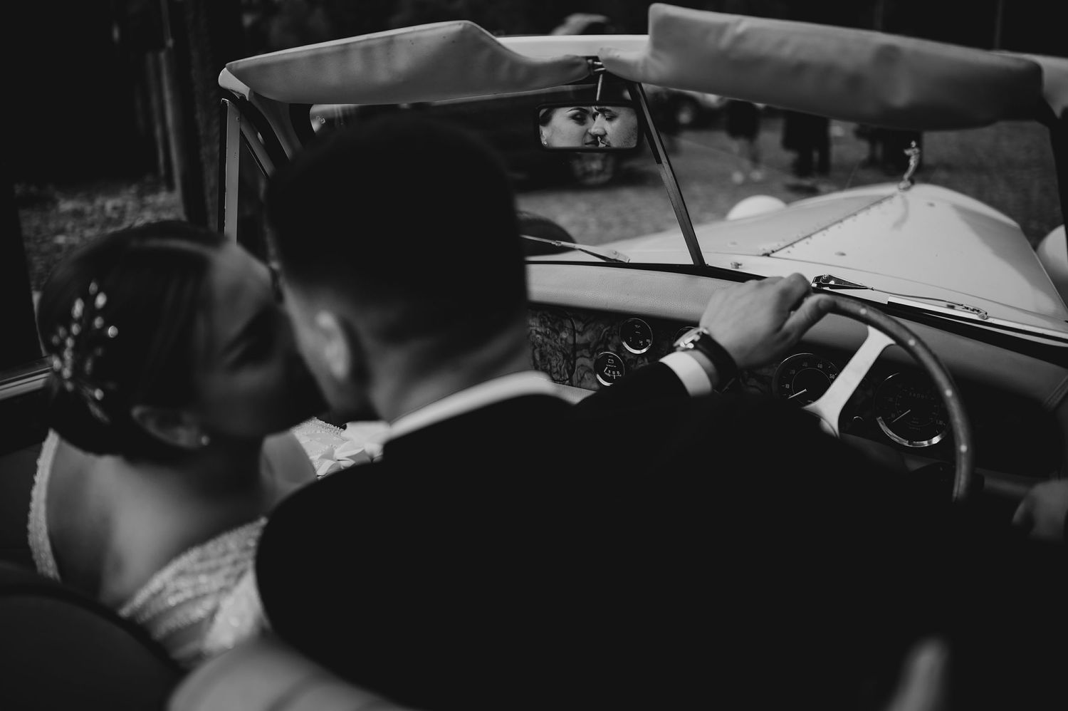 Black and white photo of bride and groom kissing inside a vintage convertible wedding car.
