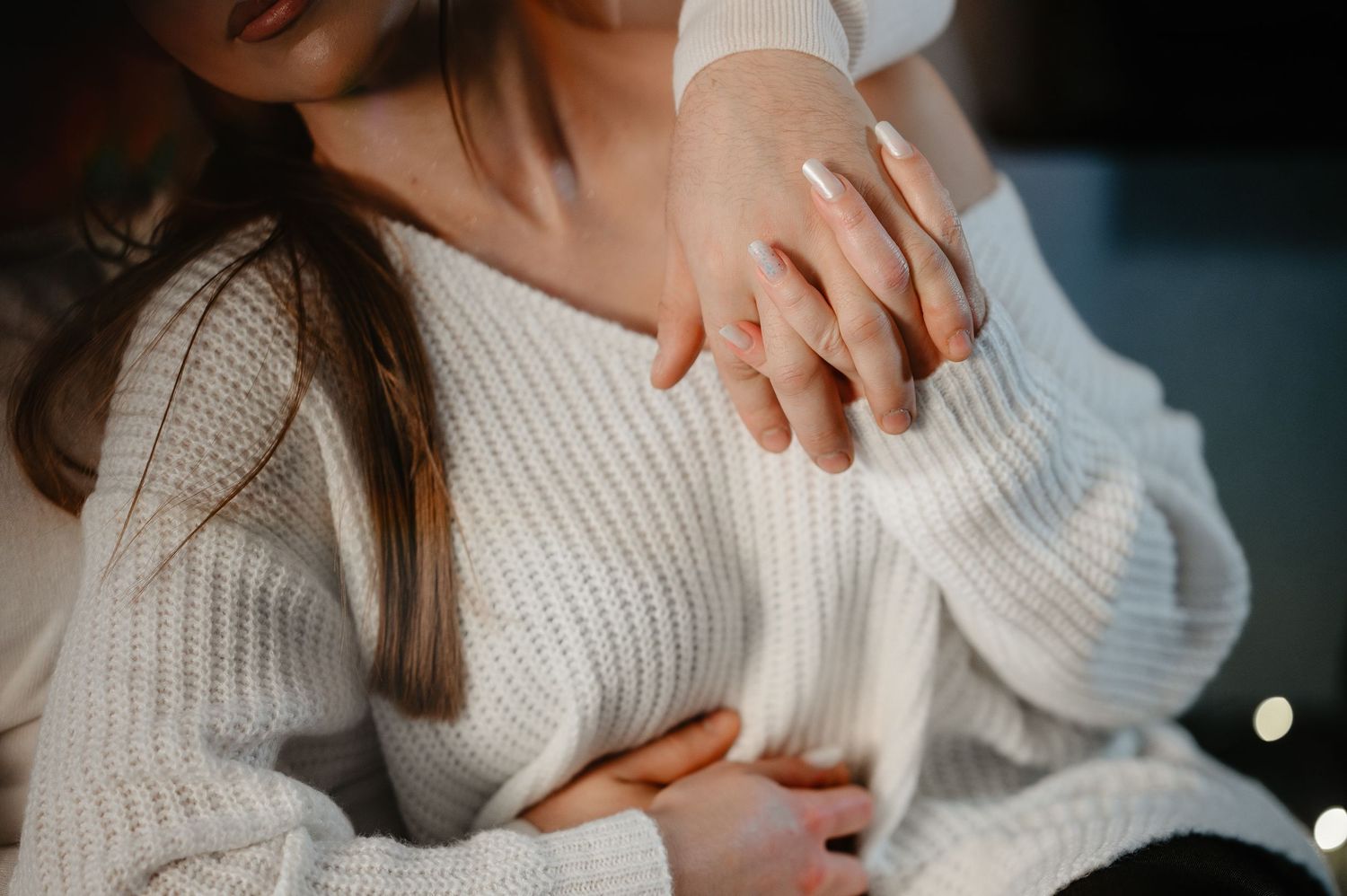 Close-up of woman in white knit sweater with manicured hands resting on her chest.