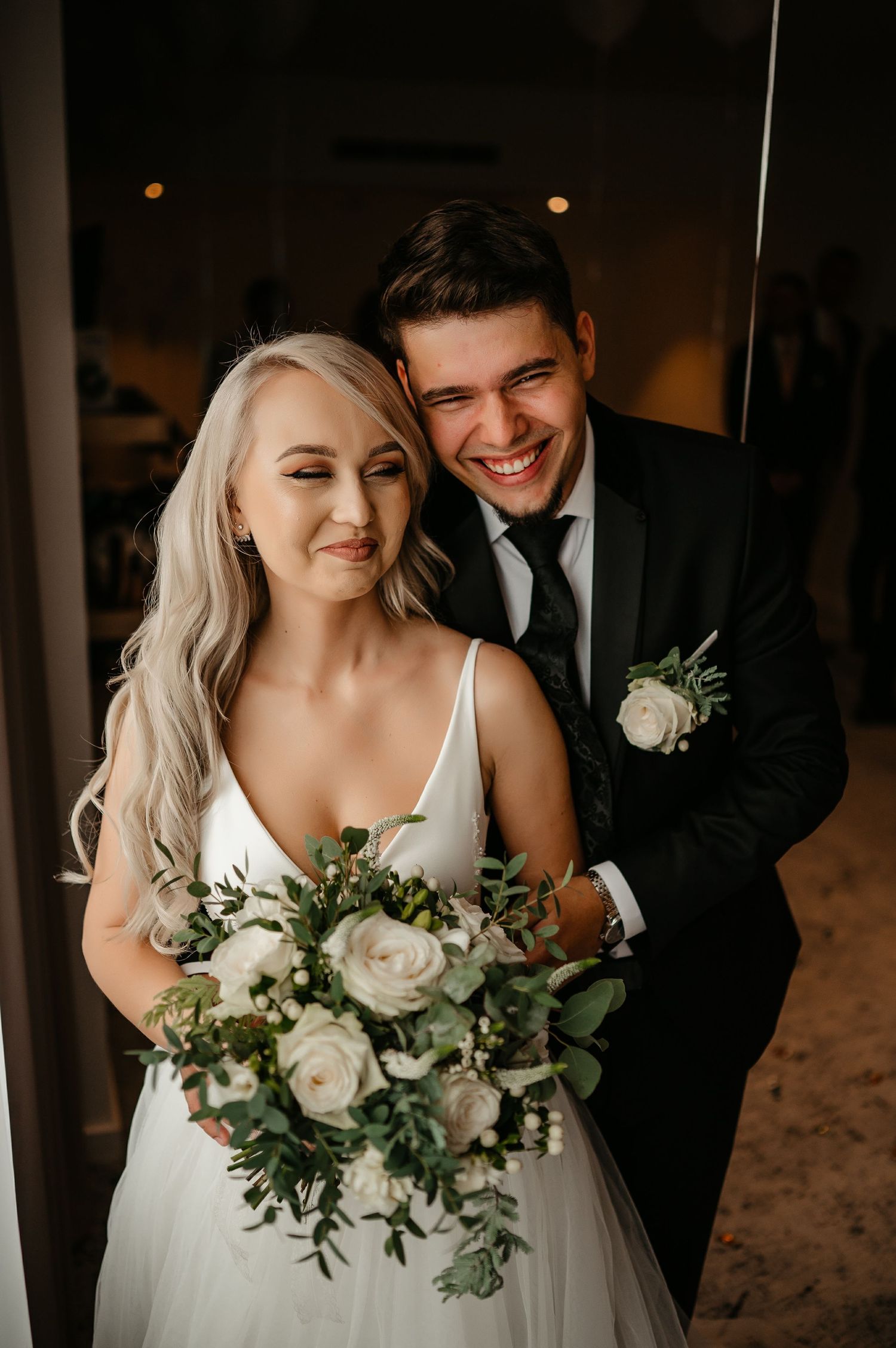Happy groom hugging blonde bride from behind as she holds white rose and greenery bouquet.