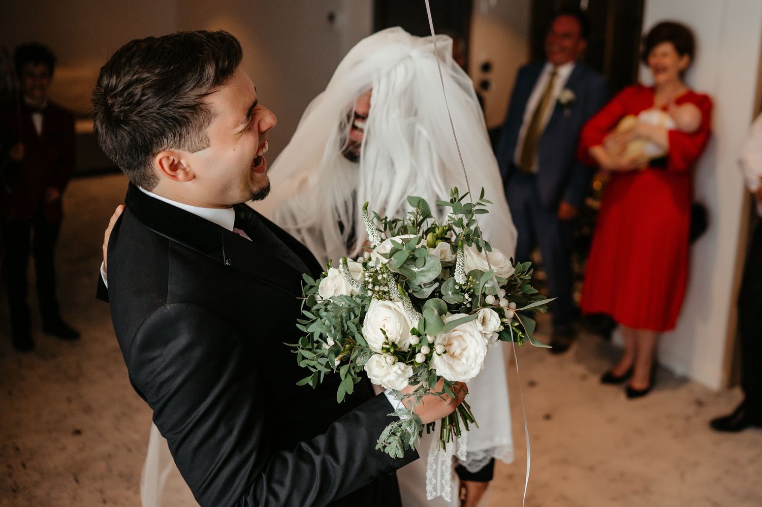 Groom embracing bride in wedding veil holding white rose bouquet with greenery at indoor ceremony.