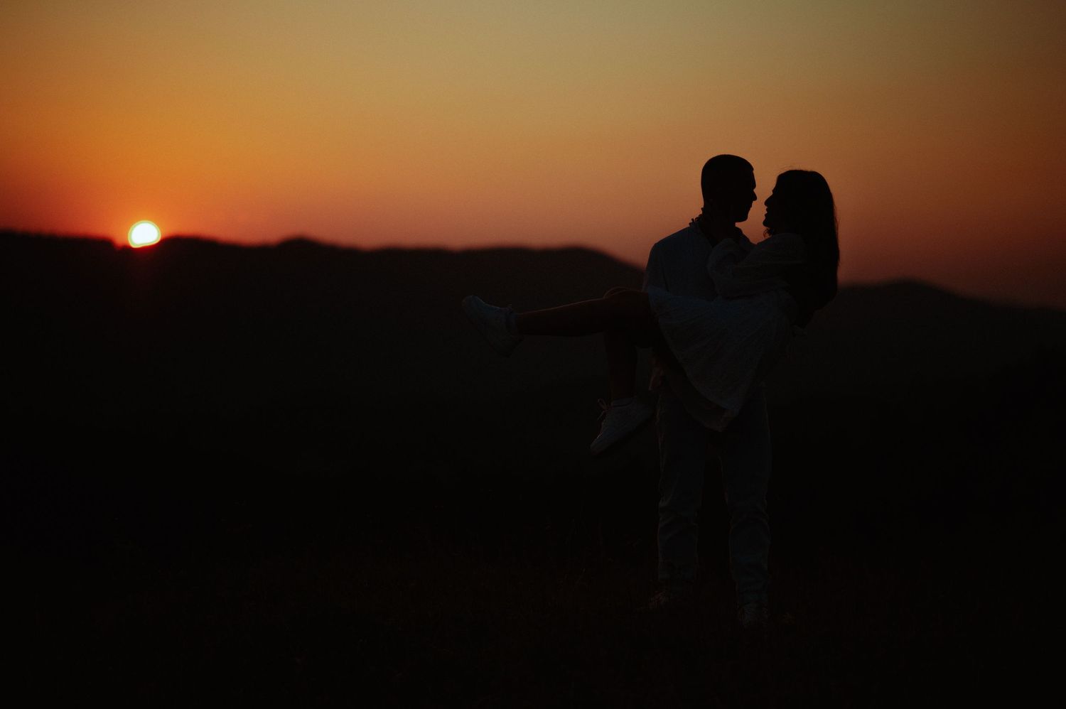 Silhouette of couple embracing at deep orange sunset with mountain horizon in background.