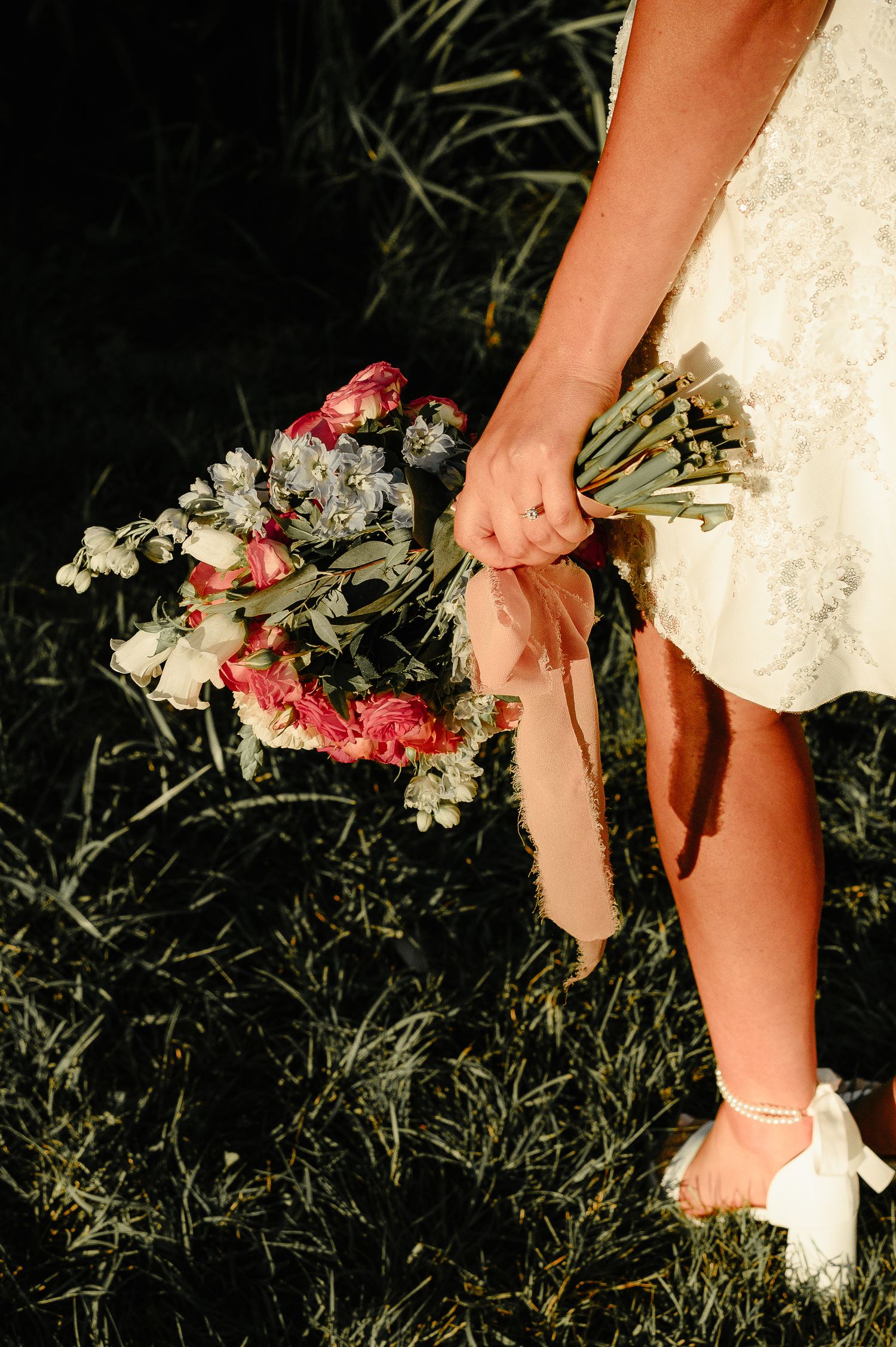 Close-up of bride holding a pink and white wedding bouquet with ribbon in a lush outdoor setting.