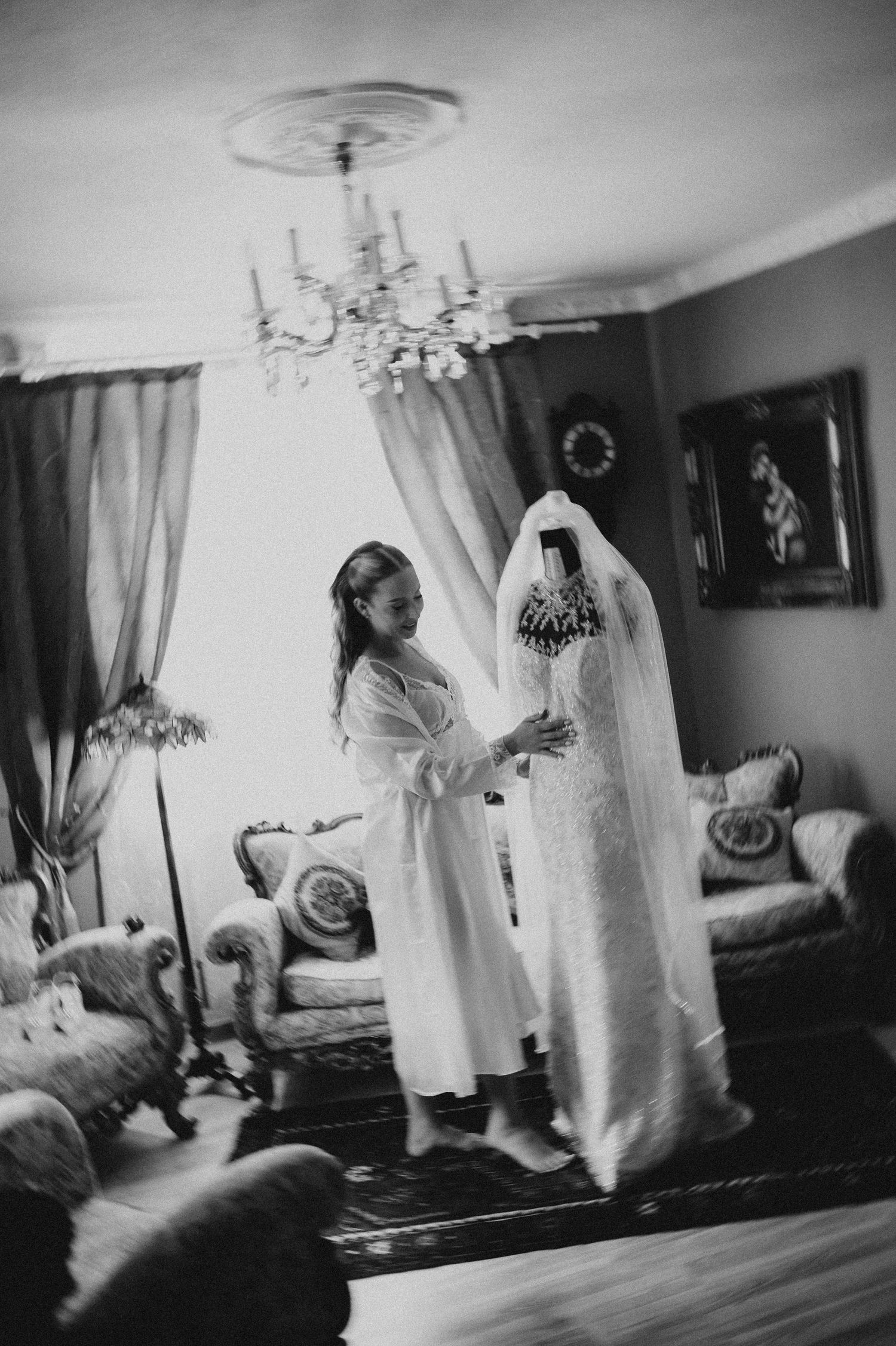 Black and white photo of a bride being helped into her wedding dress by a bridesmaid in an elegant living room.