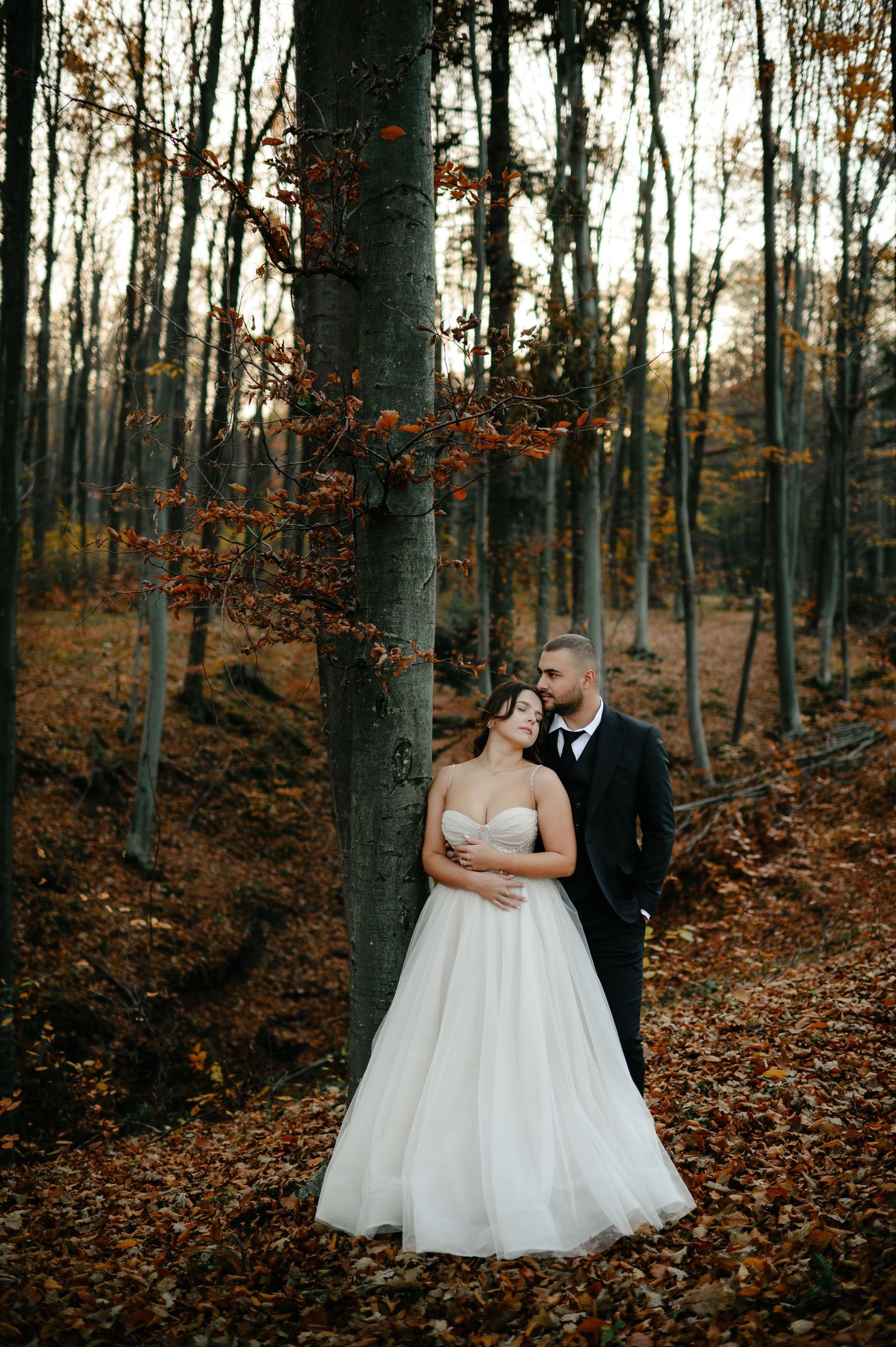 Bride and groom embracing in autumn forest, surrounded by fallen leaves and bare trees, wearing wedding attire.