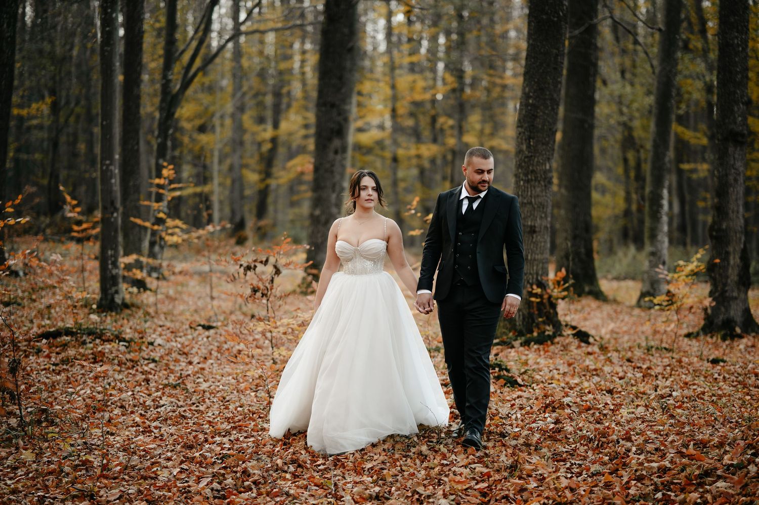 Bride and groom walk hand in hand through an autumn forest covered in fallen leaves.