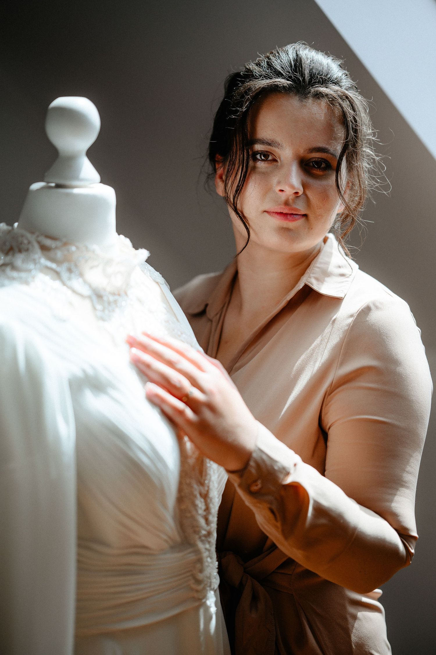 Bride in beige robe touching elegant white lace wedding dress on mannequin.