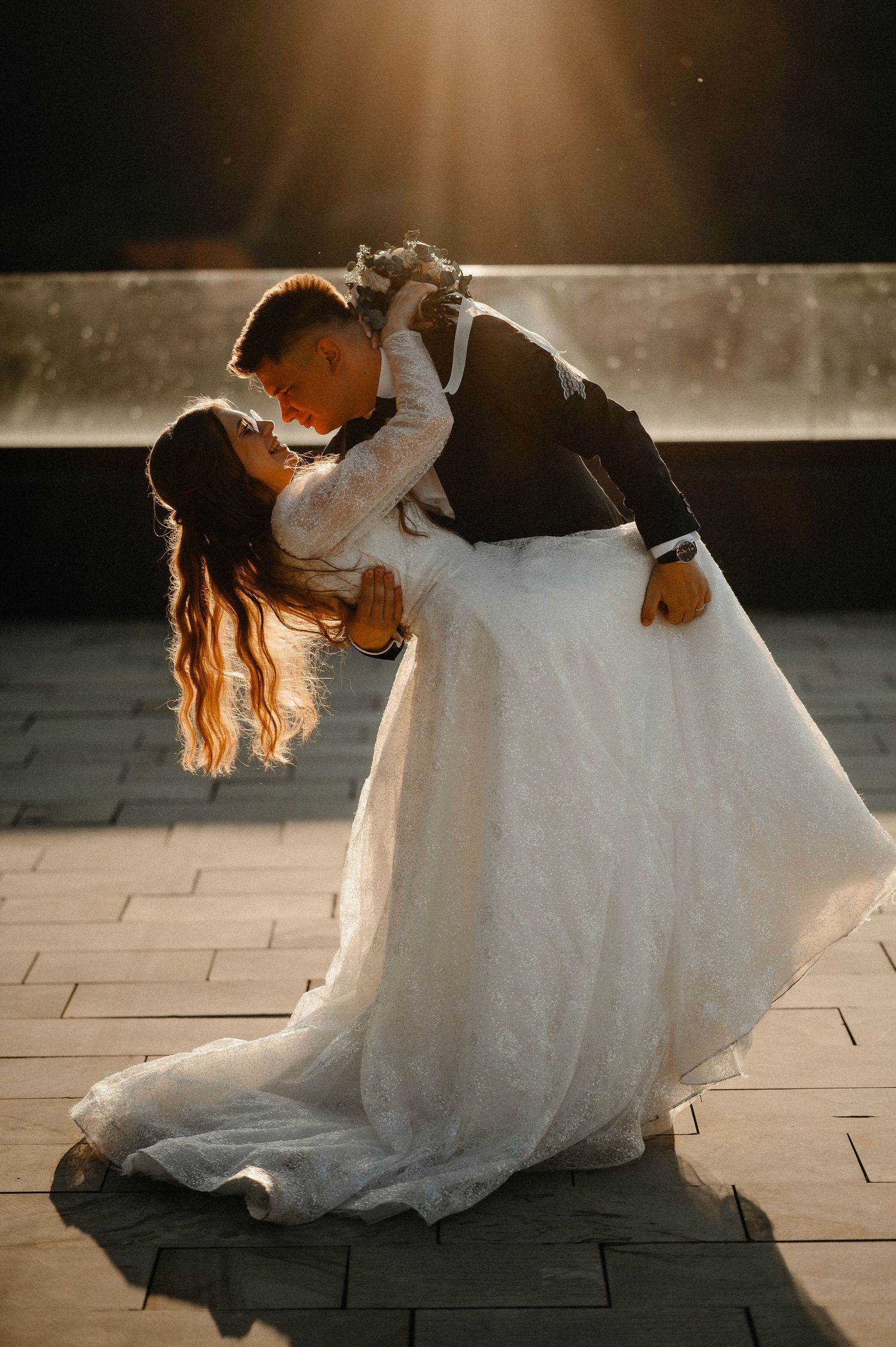 Bride in white gown and groom in black suit share a romantic dip kiss bathed in golden sunlight.