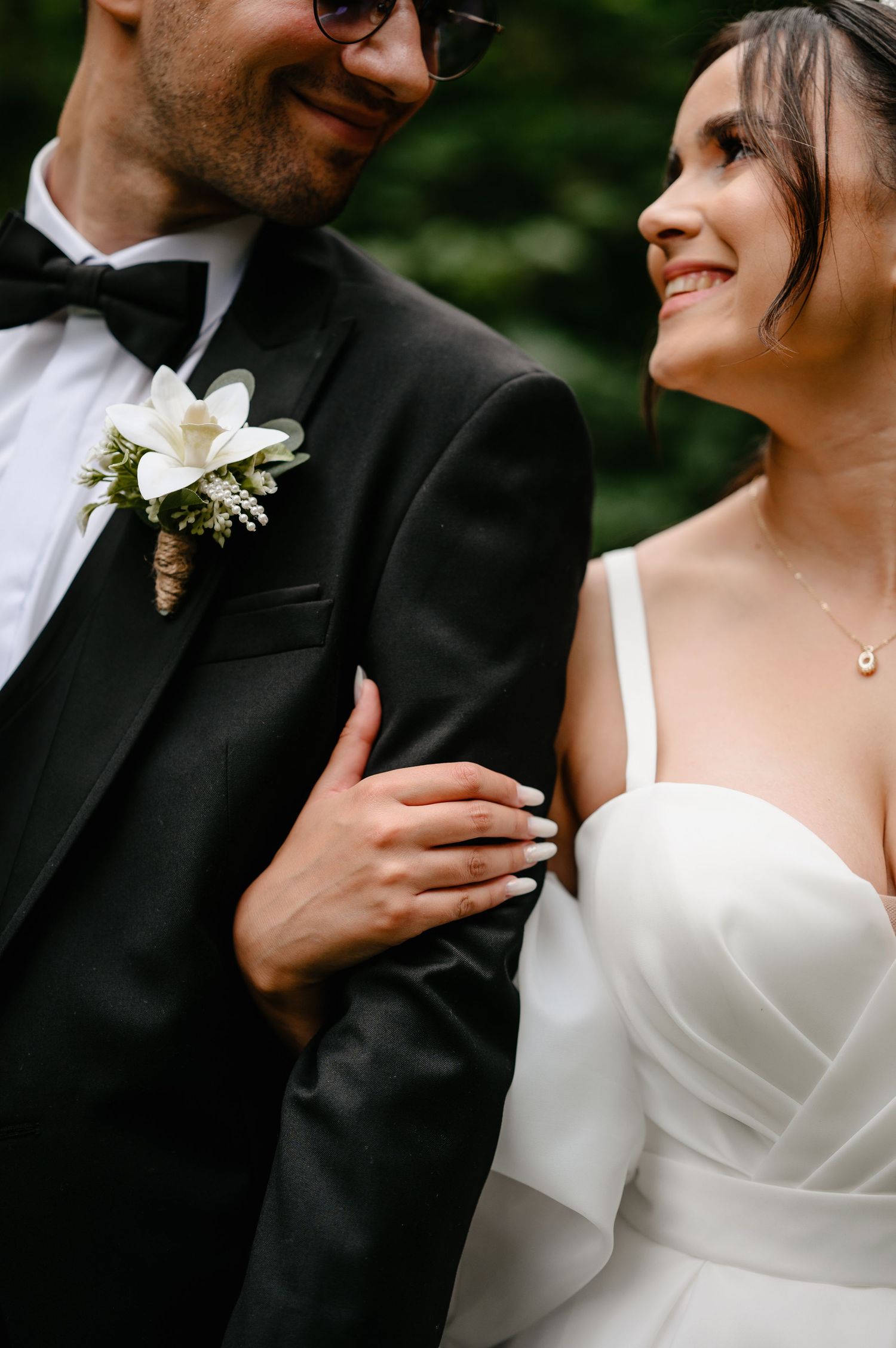 Bride in white wedding dress holds grooms arm, both smiling, with floral boutonniere detail visible.
