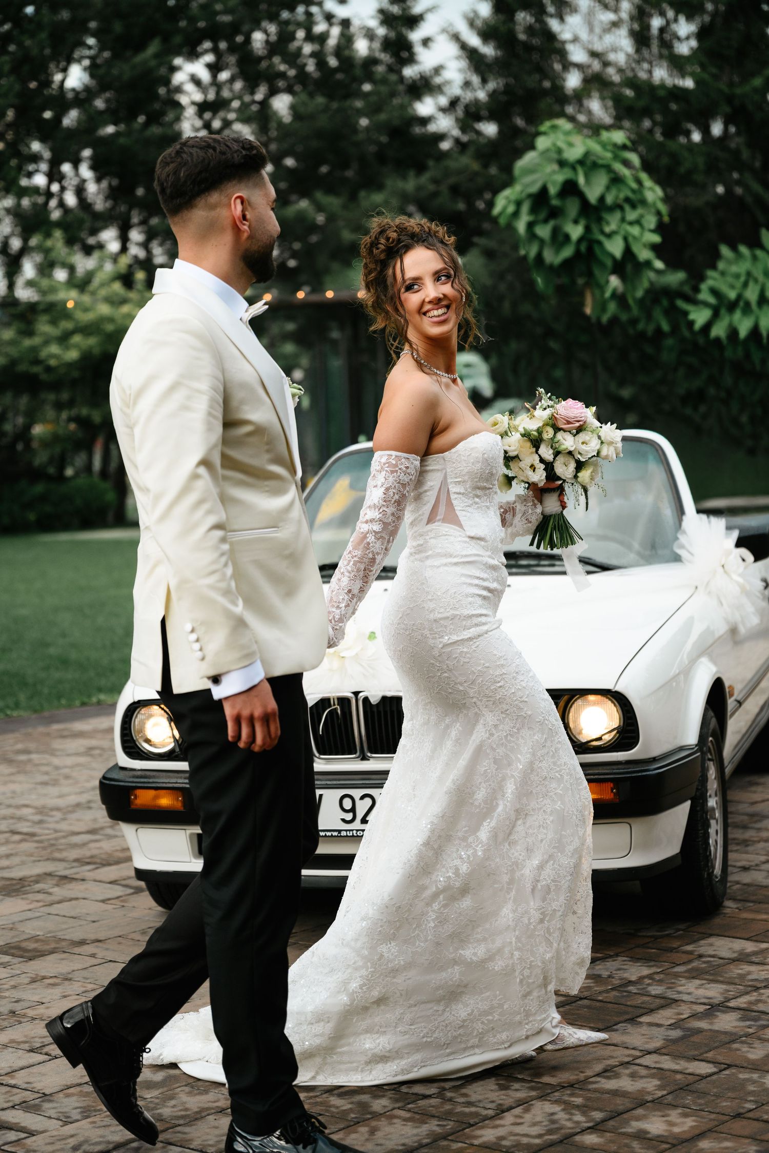 A smiling bride in a lace gown holds a bouquet beside a white BMW convertible, facing her groom in a white tuxedo jacket.