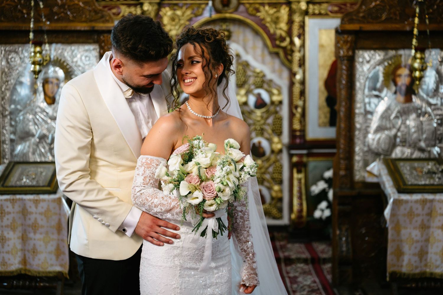 Bride and groom share a romantic moment in an ornate Orthodox church, bride holding a white and pink bouquet.