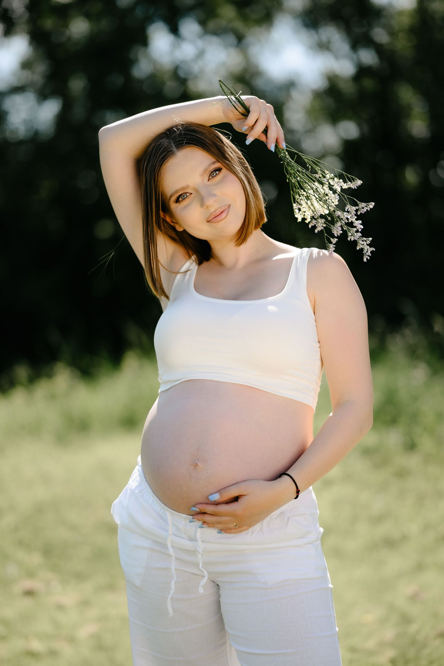 Pregnant woman in white crop top and pants holding wildflowers outdoors in sunlit green field.
