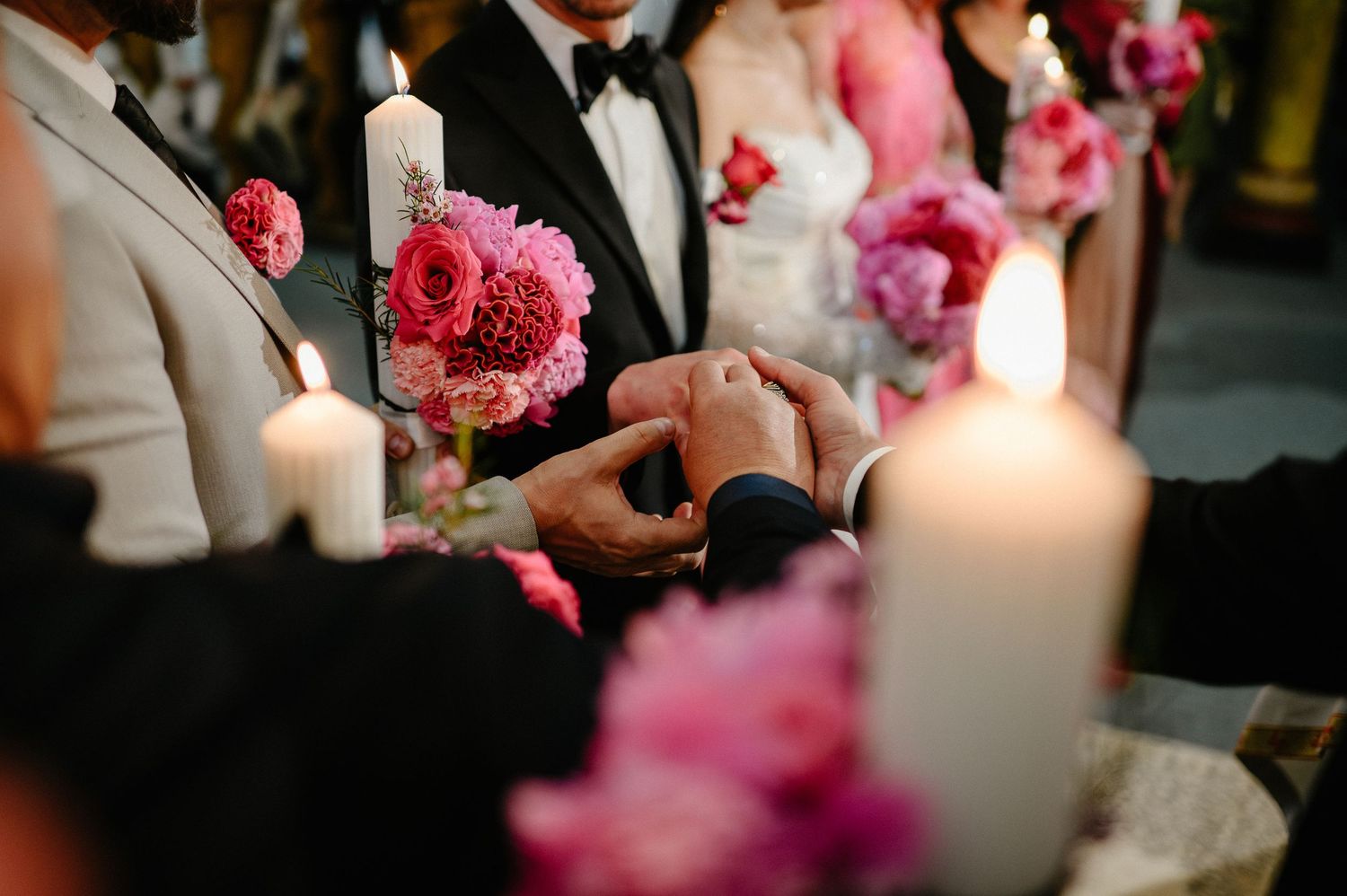 Close-up of hands exchanging rings at a candlelit wedding altar surrounded by pink peony arrangements.