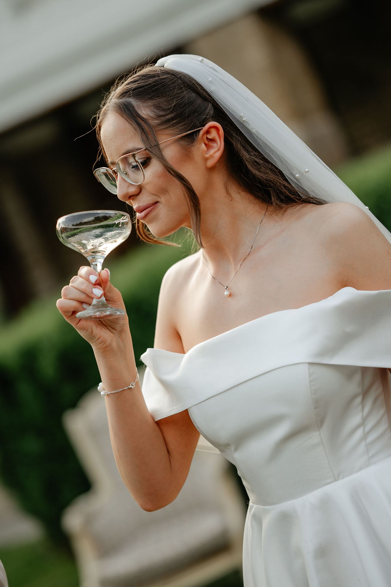 Bride in white off-shoulder gown and veil sipping champagne from a coupe glass outdoors.