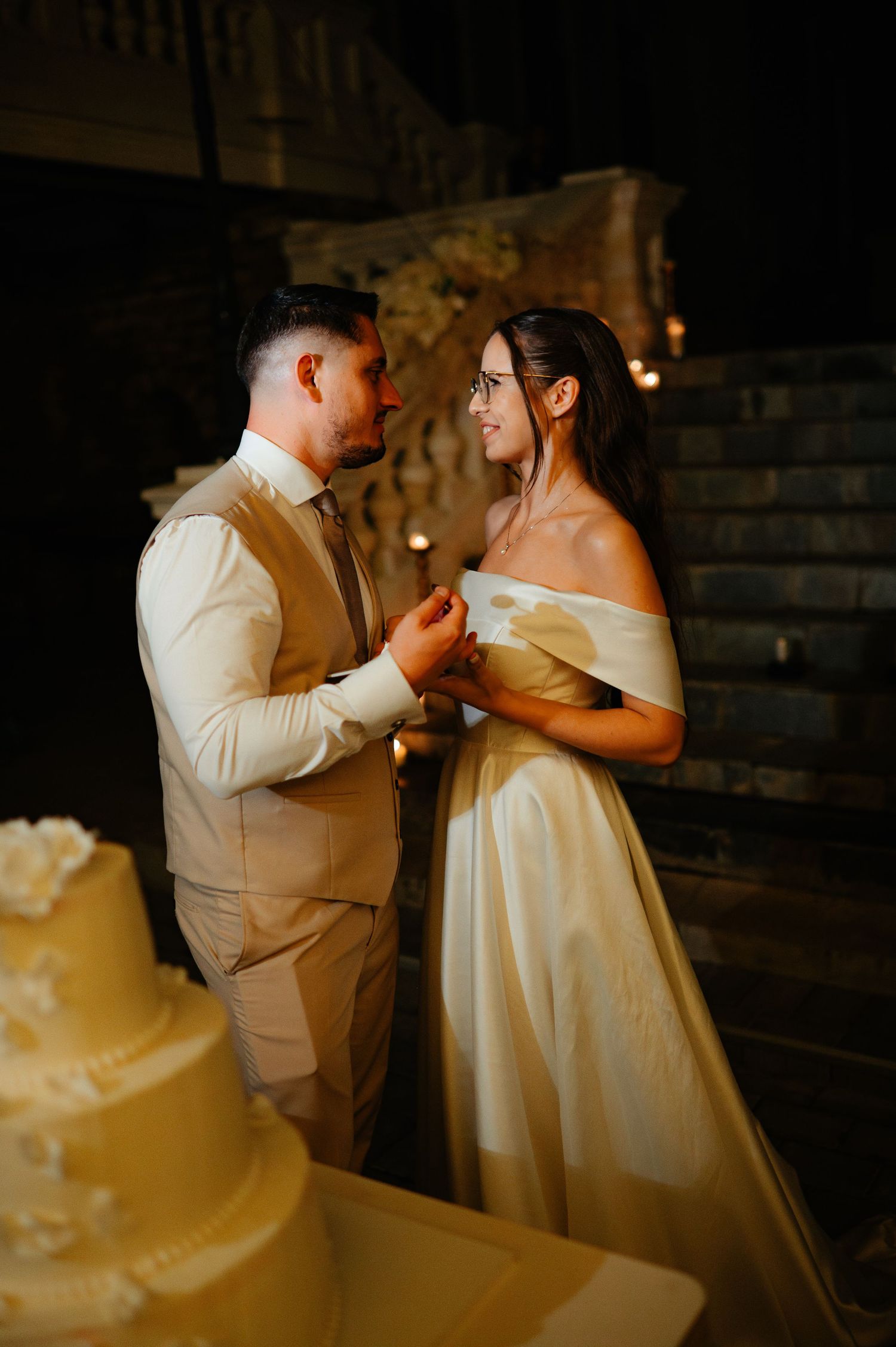 Newlyweds share a joyful moment holding hands beside white tiered cake at romantic night wedding.