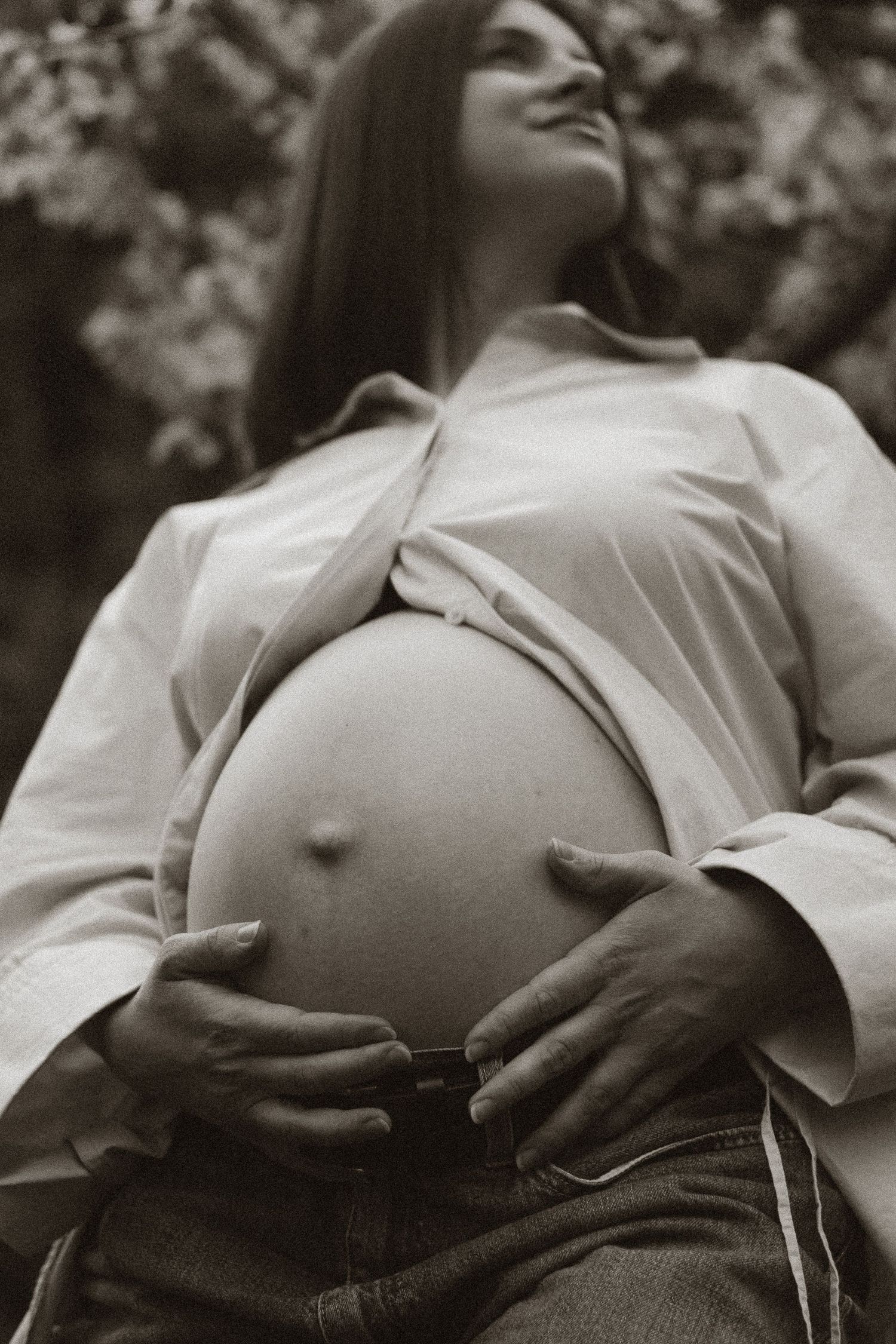 Sepia toned image of smiling pregnant woman holding baby bump in open white shirt.