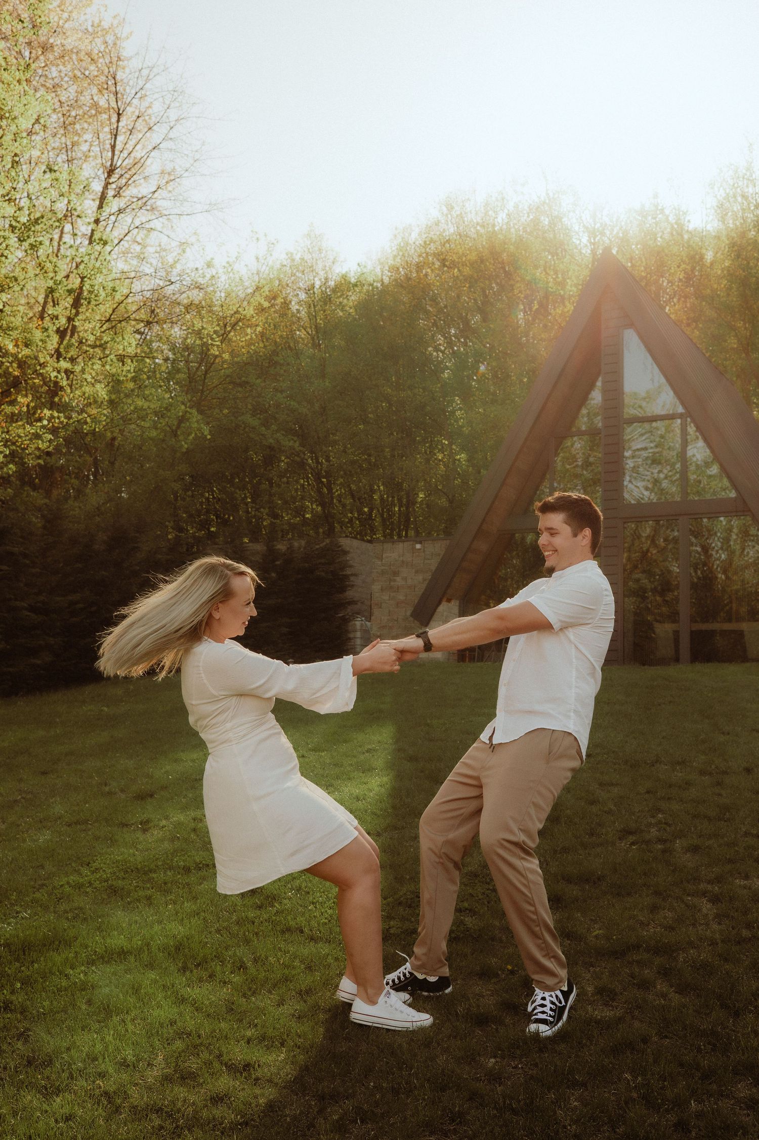 Couple dancing and spinning together on green grass in golden hour sunlight near an A-frame cabin.