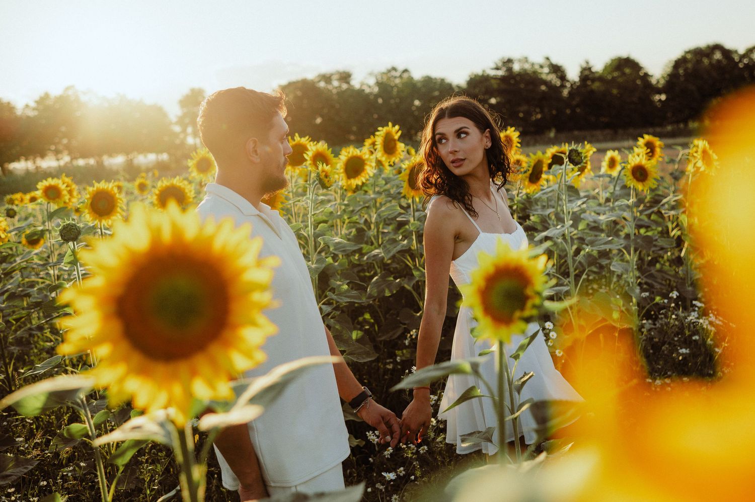 Woman in white dress looking back while holding hands with partner in sunflower field.