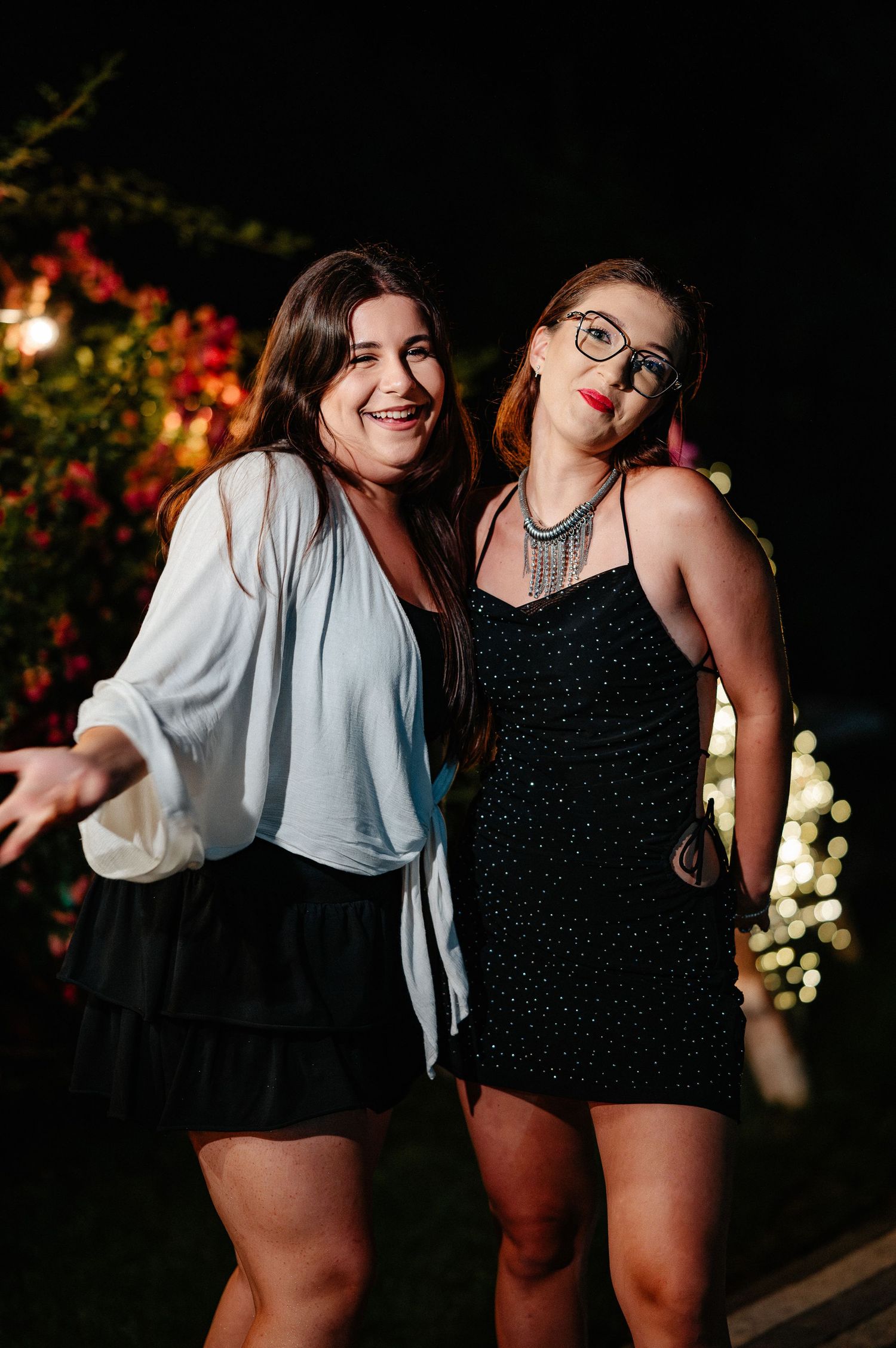 Two young women posing together at night outdoor party, one in white cardigan and one in black sparkly dress.