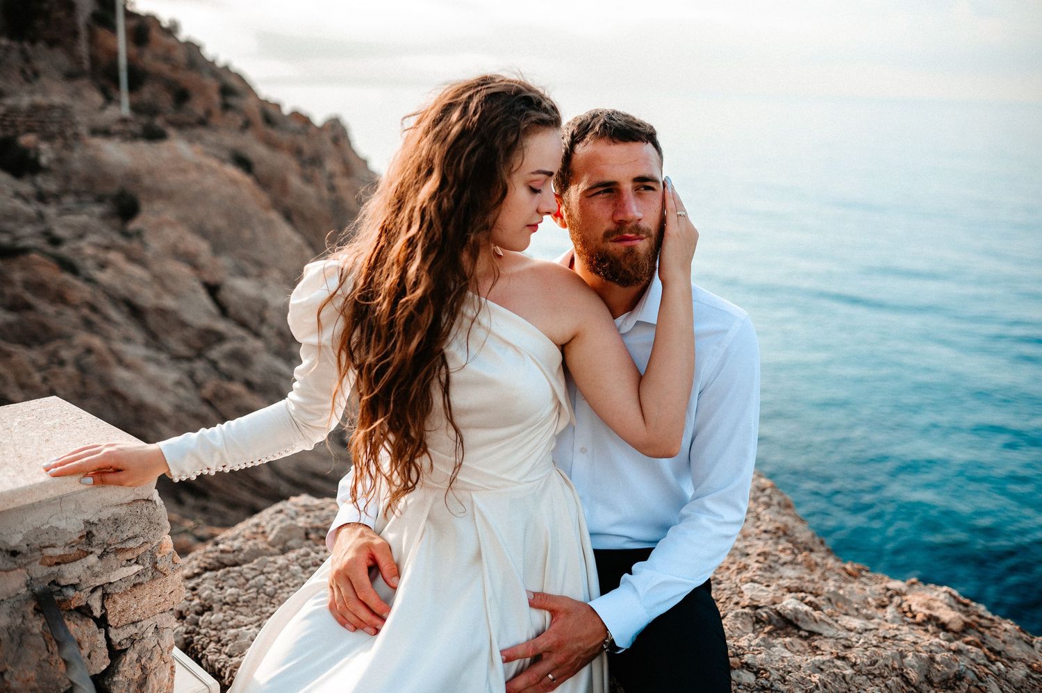 Romantic couple embracing on rocky seaside cliff, woman in white wedding dress with ocean view behind them.