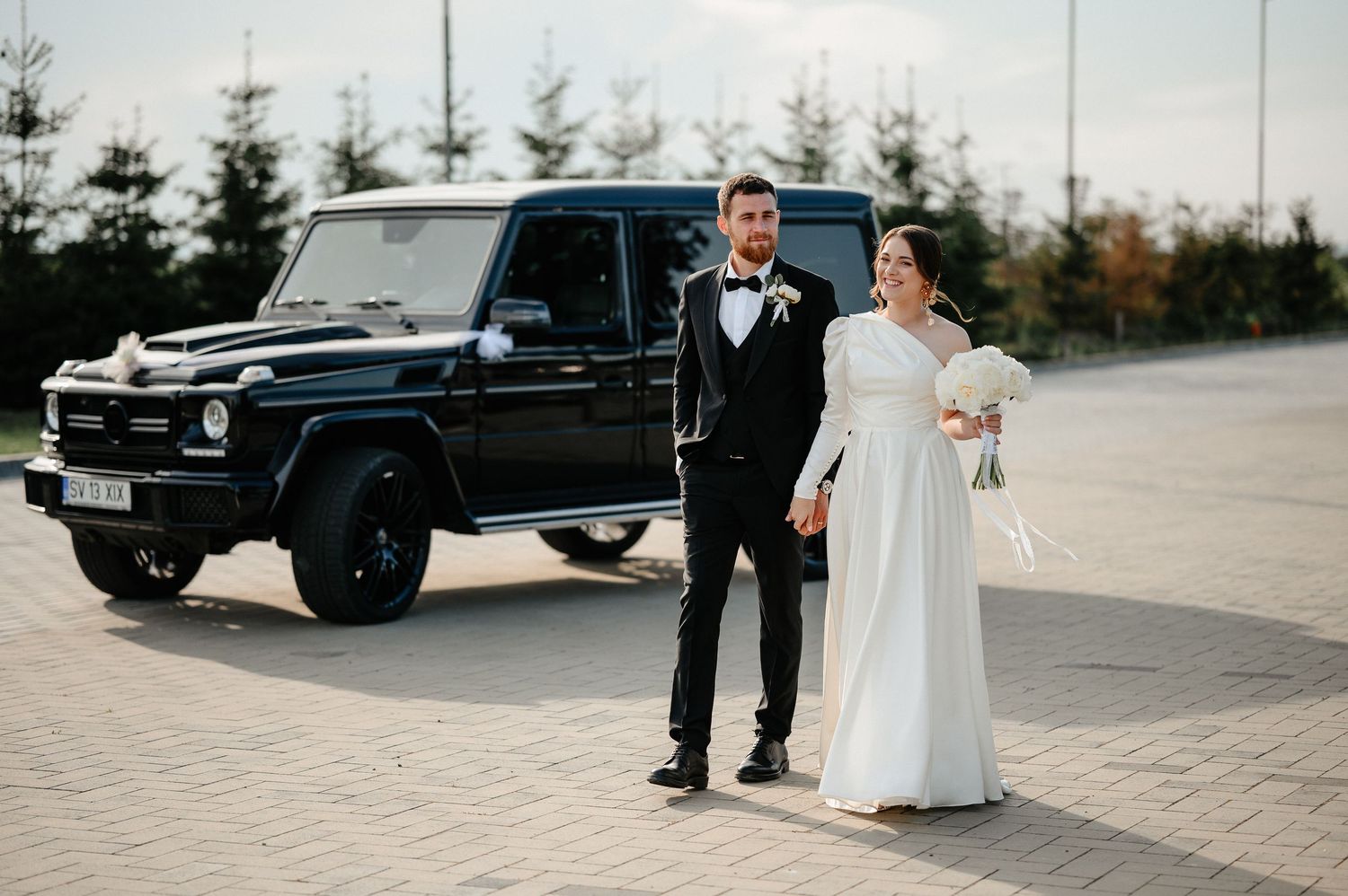 Bride and groom posing together in front of black Mercedes G-Wagon on wedding day.