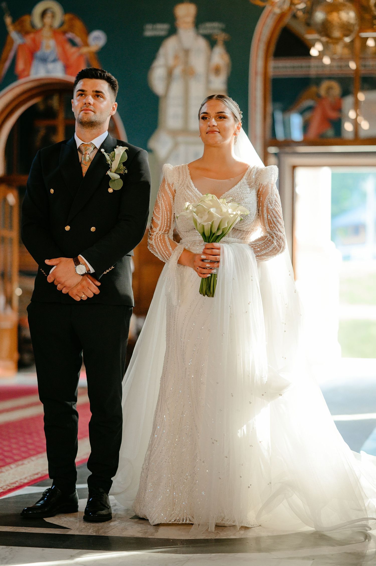 Bride and groom standing at the altar during a Orthodox church wedding ceremony, bride holding white calla lily bouquet.
