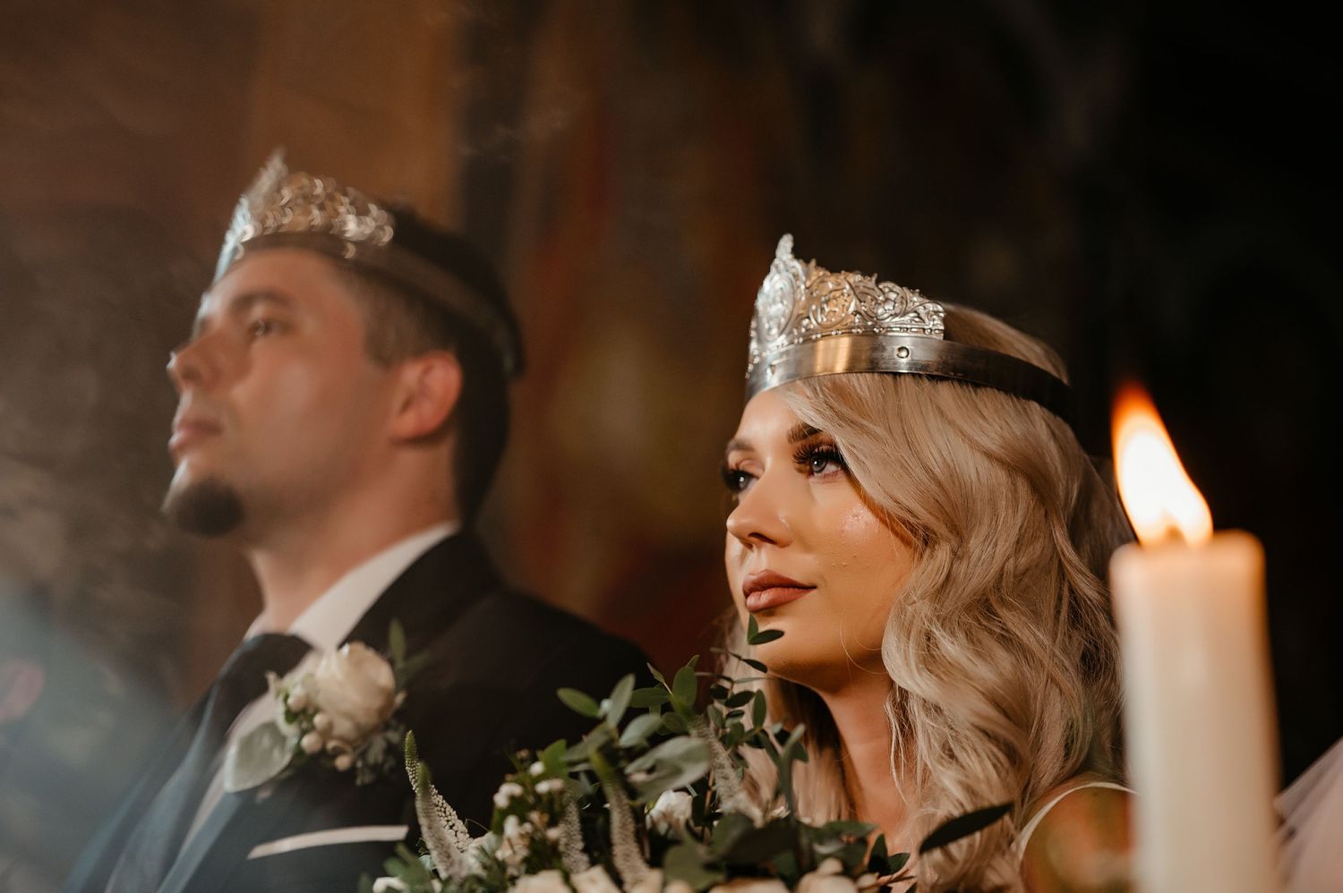 Bride and groom wearing crowns during Orthodox wedding ceremony with candlelight.