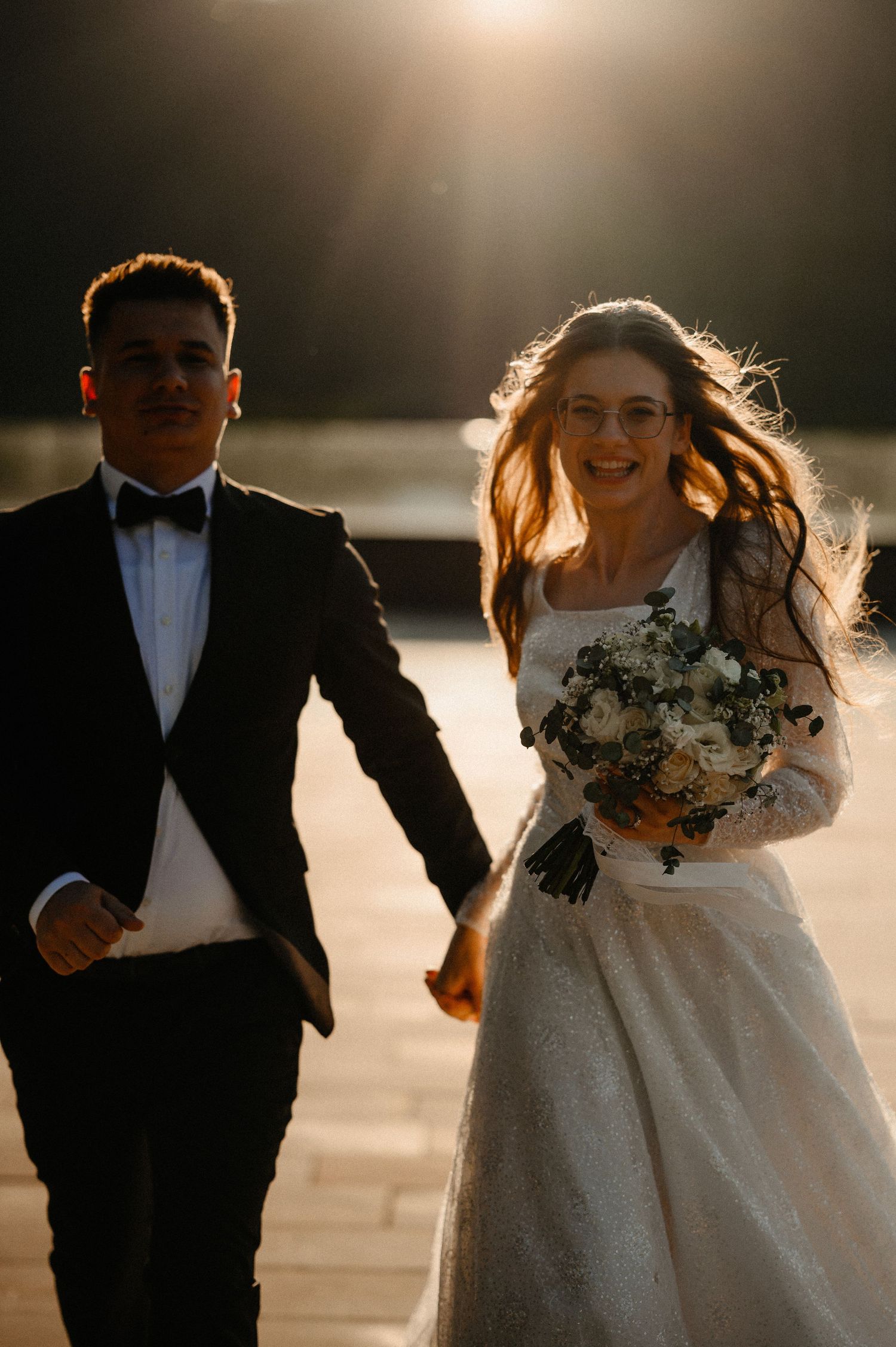 Smiling bride in white gown holding bouquet walking with groom in tuxedo in golden sunlight.