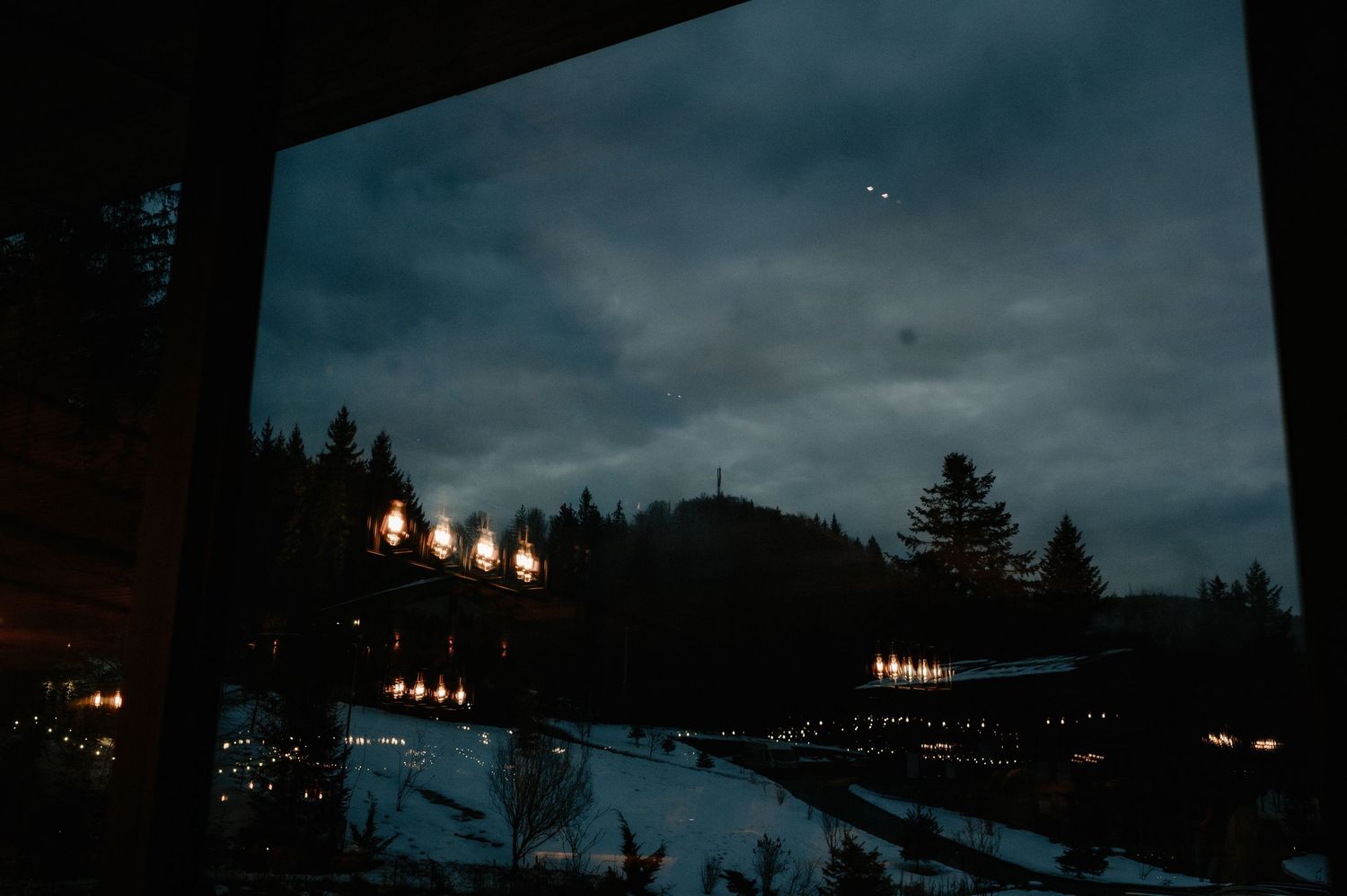 Dark winter evening landscape view through window showing snow-covered mountains with glowing lights.
