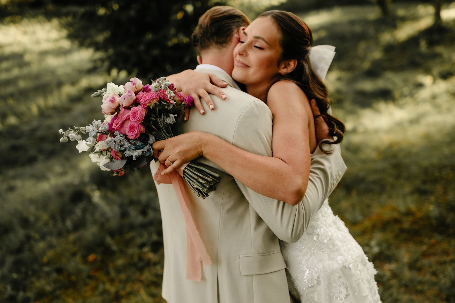 Smiling bride hugs groom tightly outdoors, holding a beautiful pink floral bouquet on their wedding day.