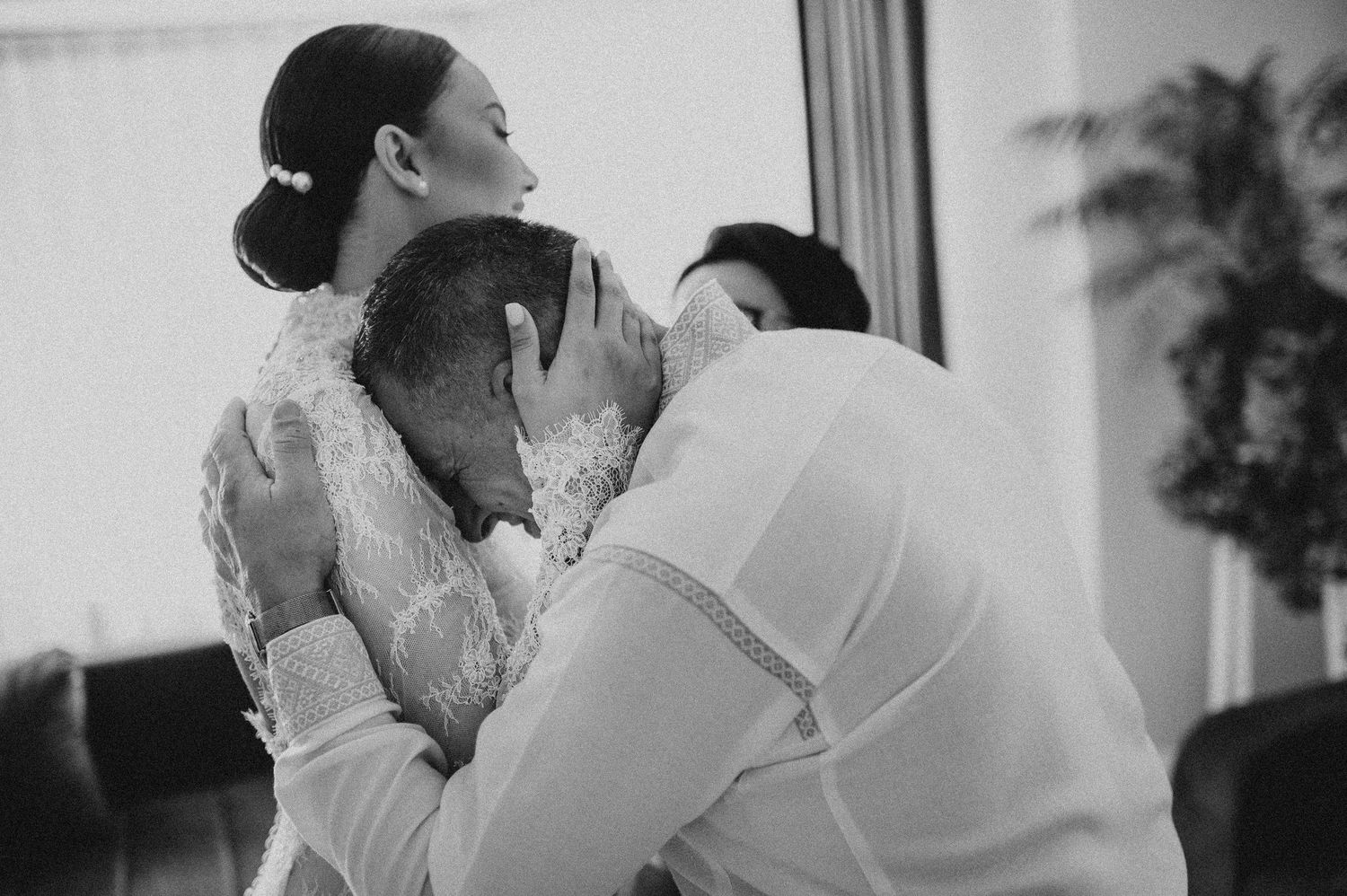 Tender black and white moment of an emotional family member bowing head while embracing the lace-gowned bride.