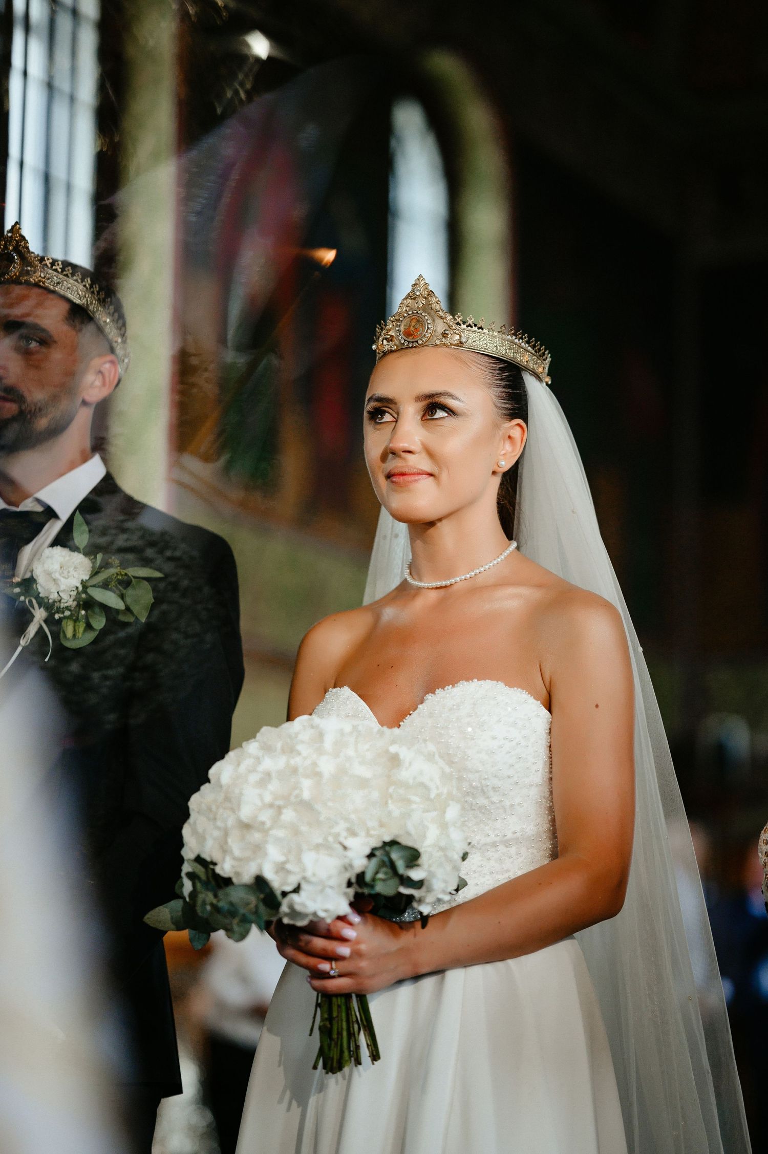 Radiant bride wearing golden crown and veil holds white hydrangea bouquet during Orthodox wedding ceremony.