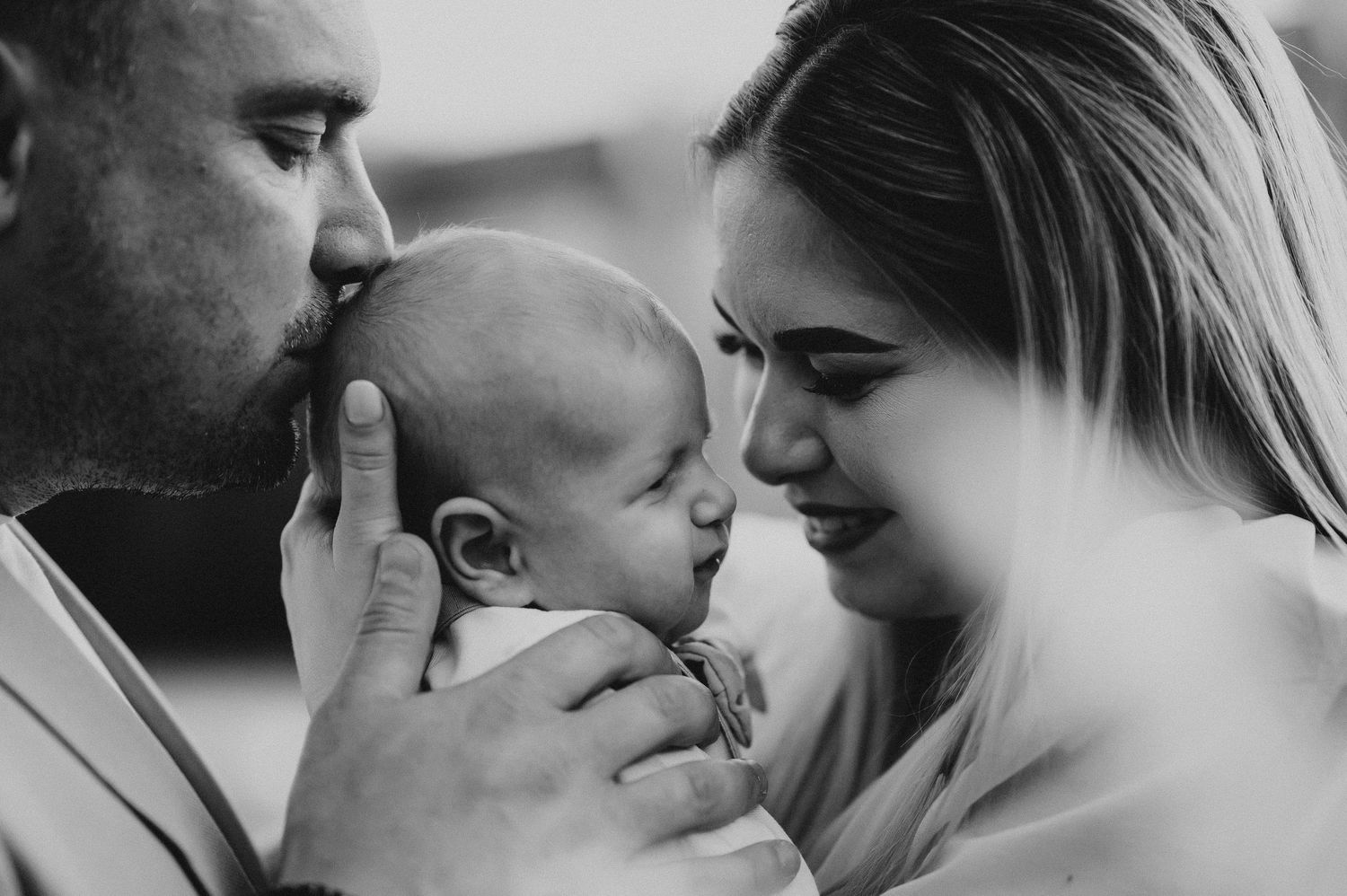 Black and white close-up of parents tenderly kissing and gazing at their sleeping newborn baby.