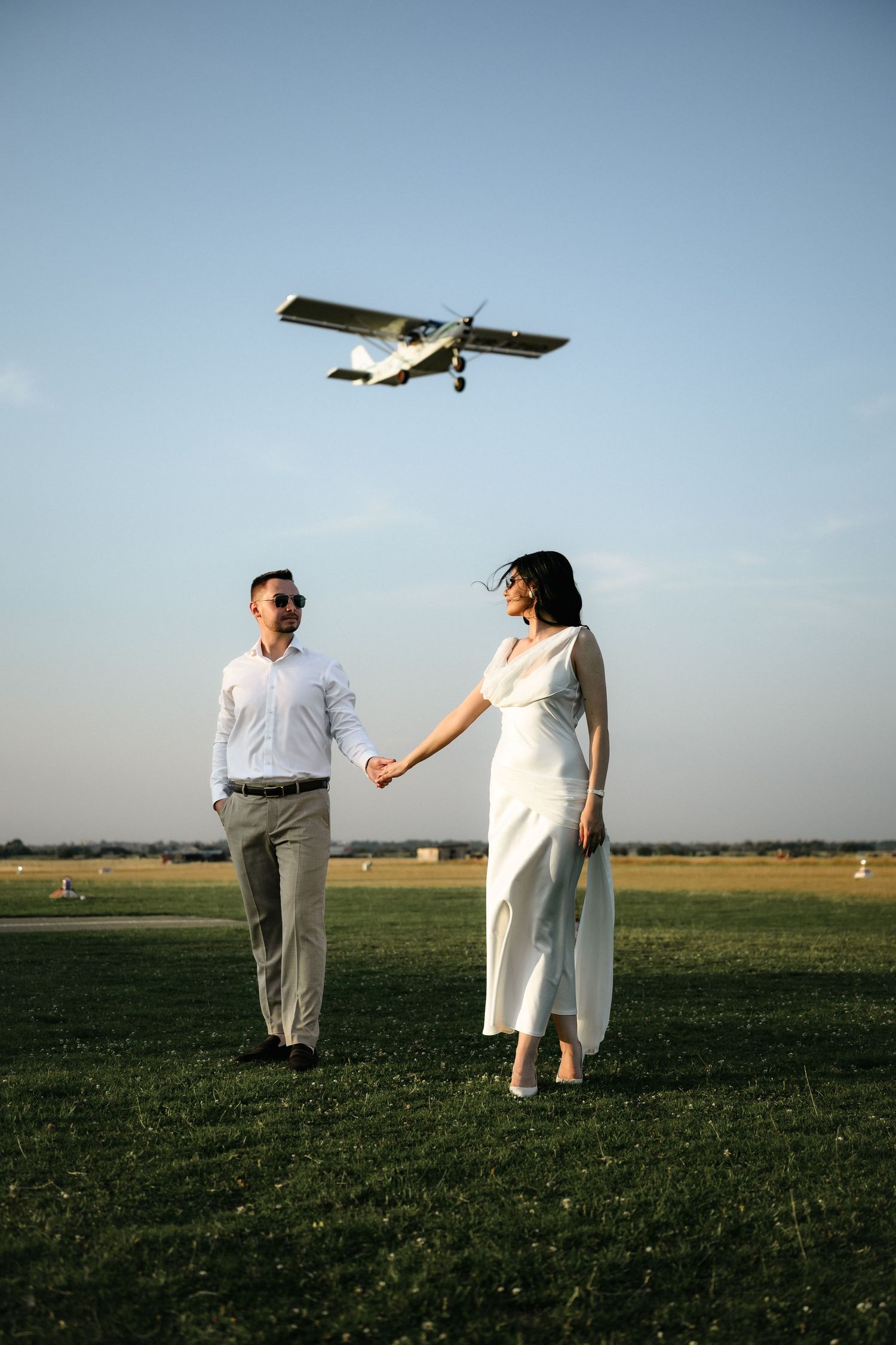 Couple holding hands on green airfield with small plane flying overhead in clear blue sky.