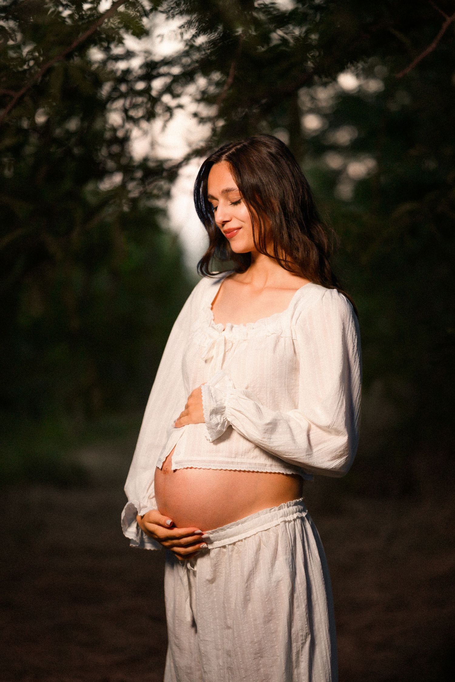 Pregnant woman in white boho outfit cradling her baby bump outdoors in golden sunset light among green trees.