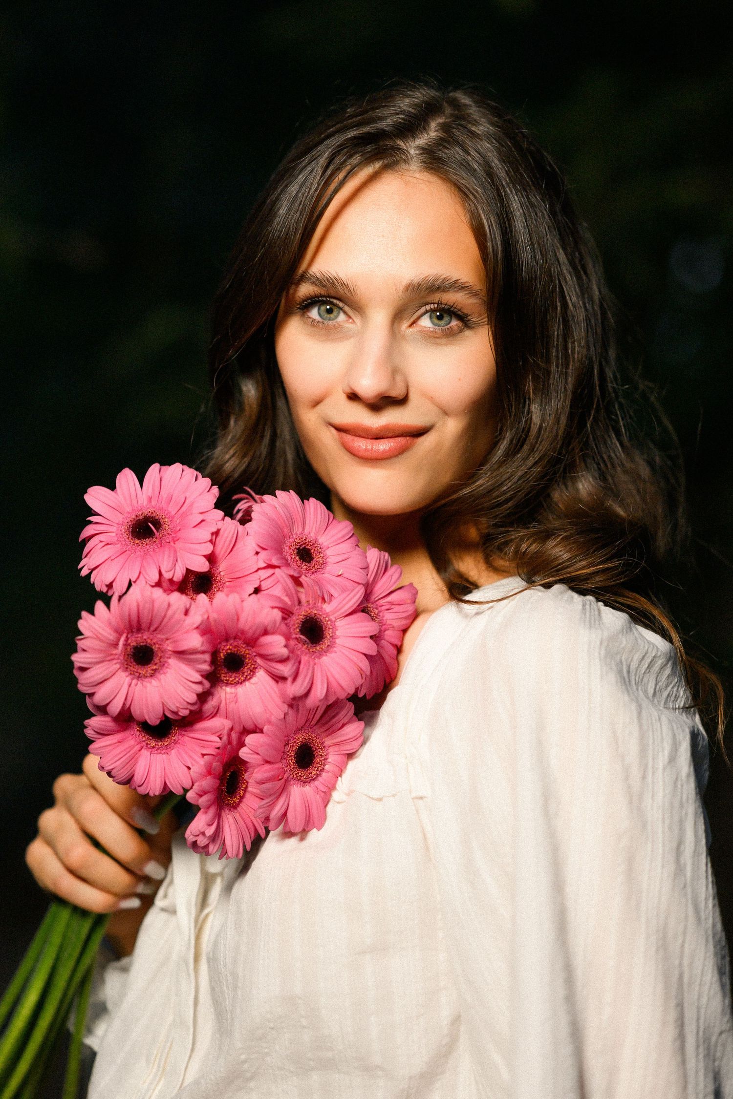 Young woman with green eyes holding a bouquet of pink gerbera daisies, wearing a white blouse outdoors.